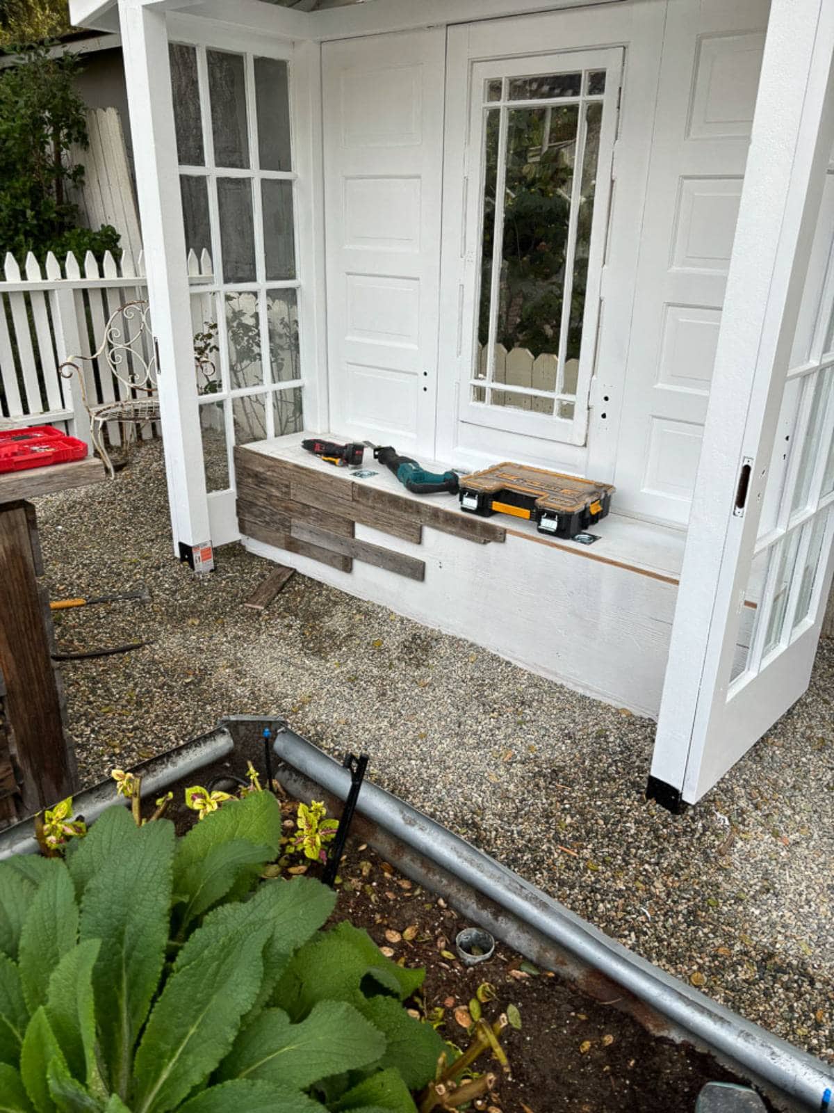 A white garden shed under construction, with wooden panels and tools on a bench. The shed has a white picket fence and a garden bed with green plants and a water pipe in the foreground. Gravel covers the ground around the shed.