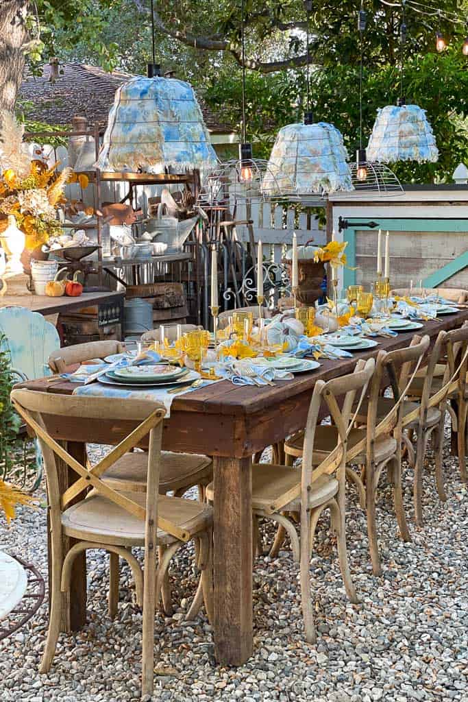 A rustic outdoor dining setup perfect for the Fall season, featuring a long wooden table with cream cloth napkins, yellow glasses, and blue-and-white dishware. Surrounding it are wooden chairs and illuminated by blue lamps, with a tree draped in a DIY rope wreath and garden elements in the background.