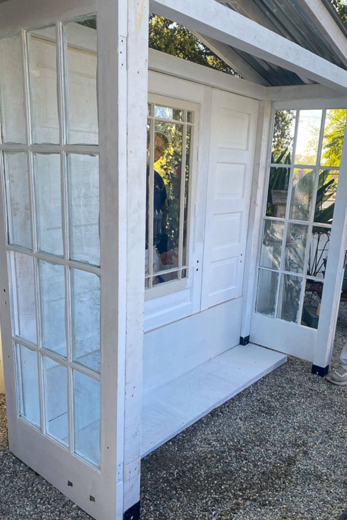 A small white shed with a door and large windows on both sides stands on a gravel surface. The shed's structure includes a transparent roof, allowing for clear sunlight to come through. There is greenery visible in the background.