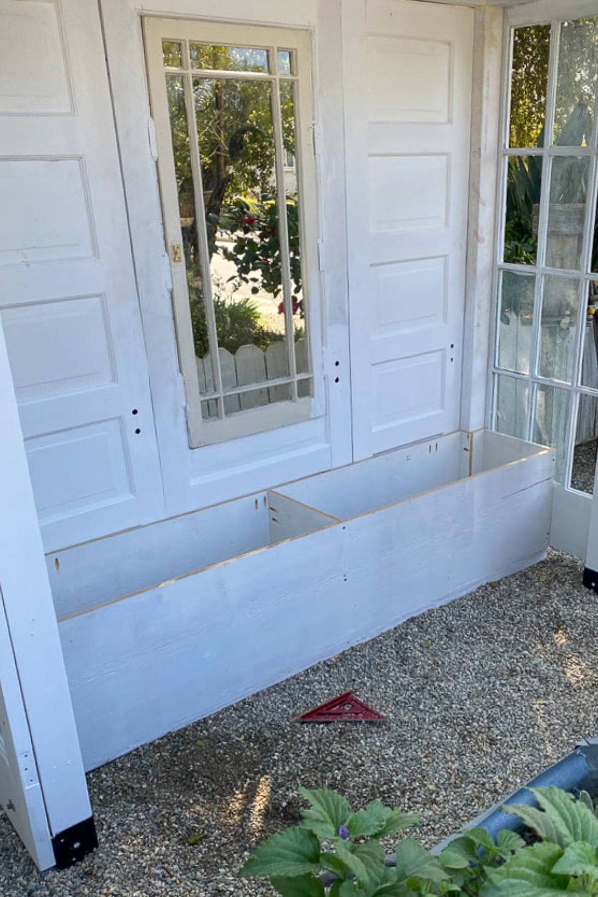 A white planter box is installed beneath a mirrored window on an outdoor white wall with surrounding white double doors. The area includes a large glass door on the right, gravel ground, and some surrounding greenery.