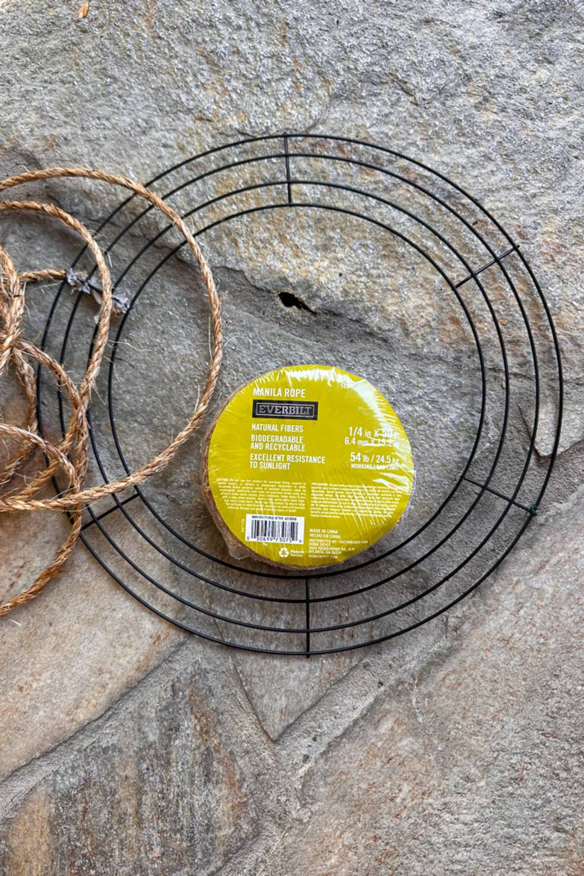 A coil of Manila rope and a black wire wreath frame are placed on a stone surface. The Manila rope is packaged in a yellow wrapper.