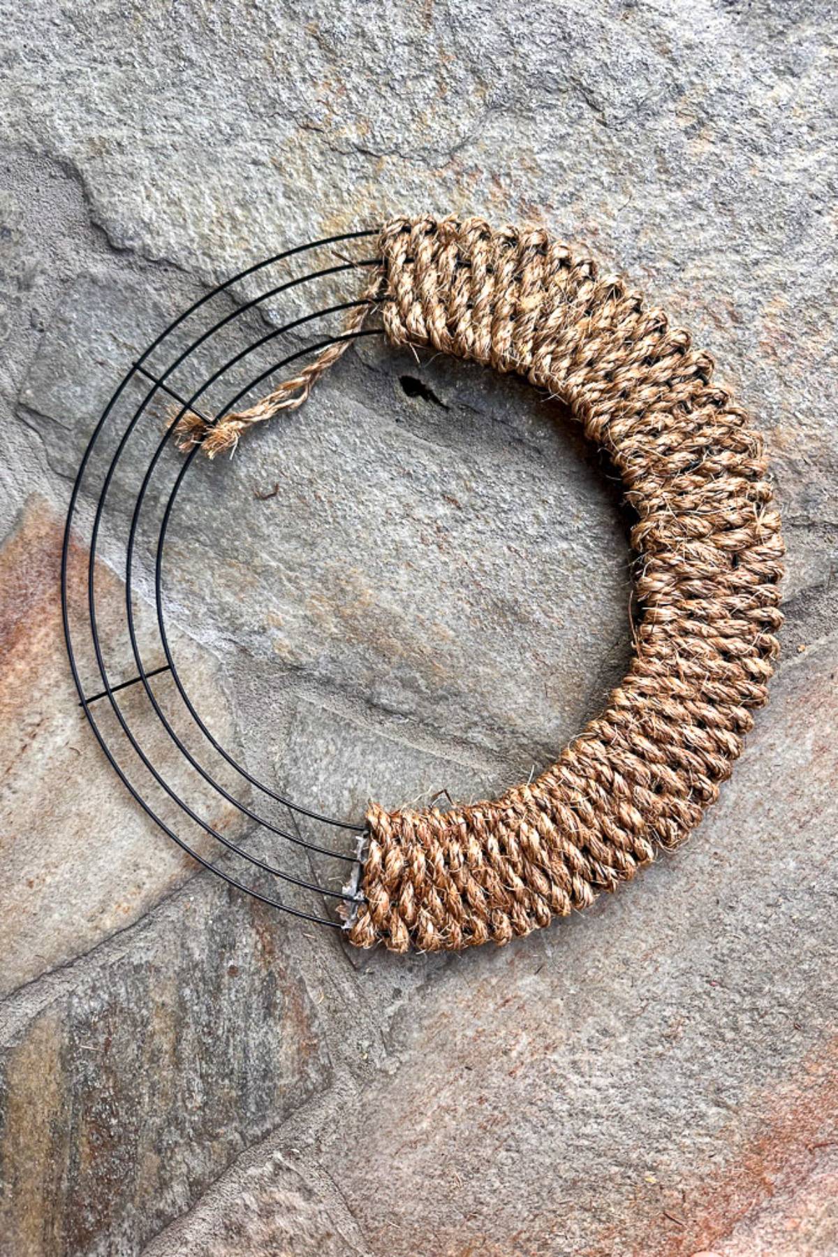 An unfinished wreath lies on a textured stone surface. The wreath is partially wrapped with brown twine, leaving part of the black metal frame exposed. The visible areas of twine are tightly wound, creating a distinct contrast against the stone background.