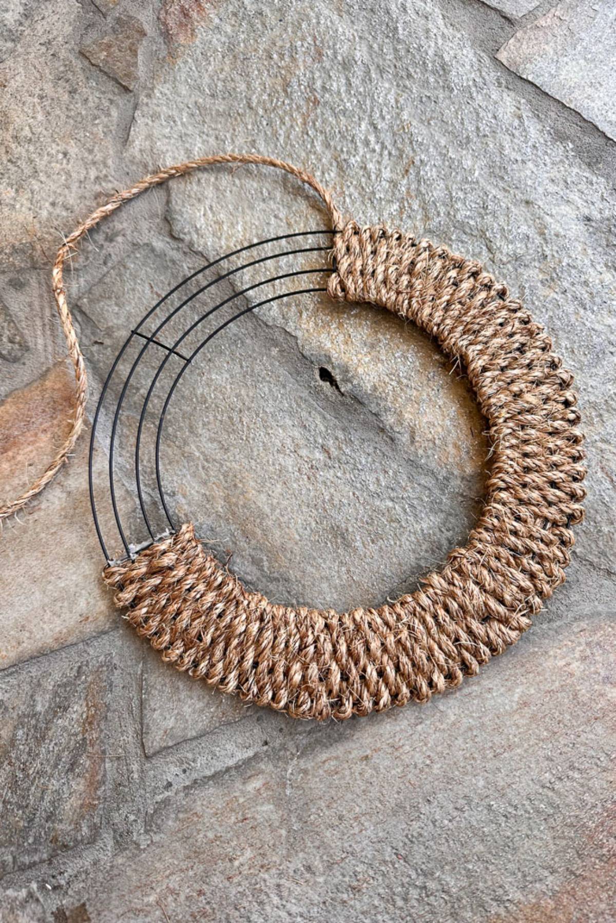 A circular pendant made of black wire partially wrapped with brown twine lies on a rough stone surface. The pendant has a rustic, handmade appearance, with the twine woven neatly around the lower part. A loose strand of twine is attached at the top.