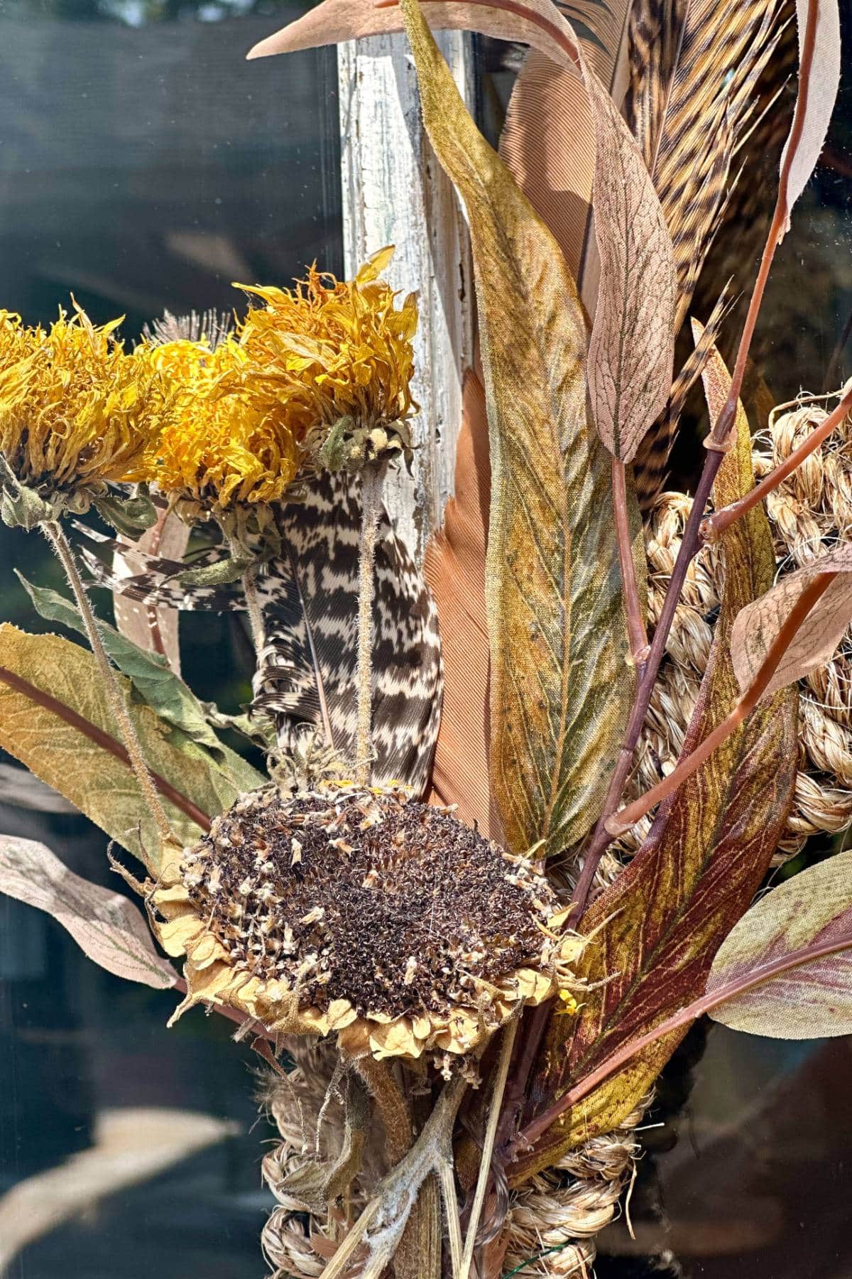 A close-up view of a dried floral arrangement featuring a variety of textures and colors. The bouquet includes sunflowers with brown centers, yellow blooms, a striped feather, and elongated leaves in shades of green and brown, all set against a reflective surface.