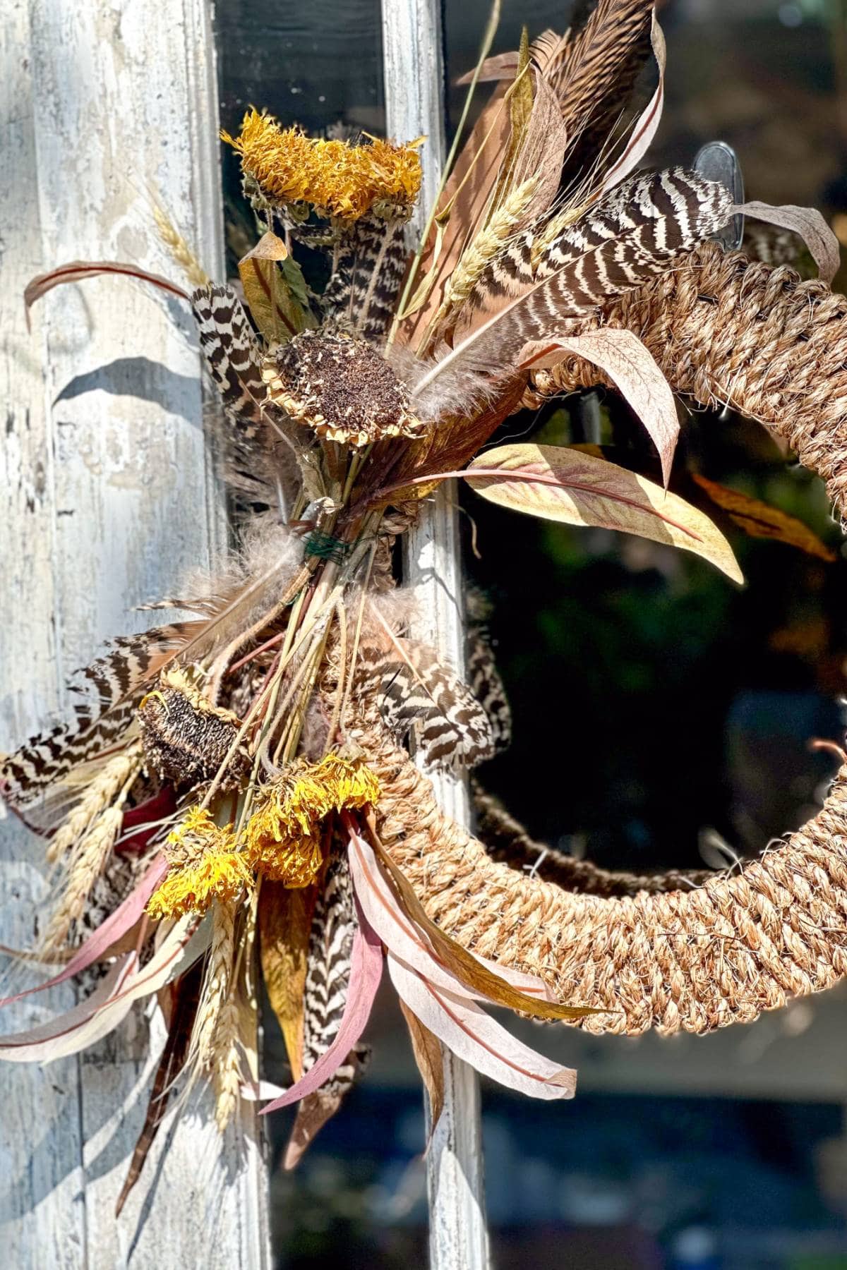 A decorative wreath made of dried grasses, feathers, and flowers hangs on a rustic wooden door. The wreath features neutral tones with accents of yellow and pink, creating an autumnal feel. The textured elements add a natural, earthy touch to the design.