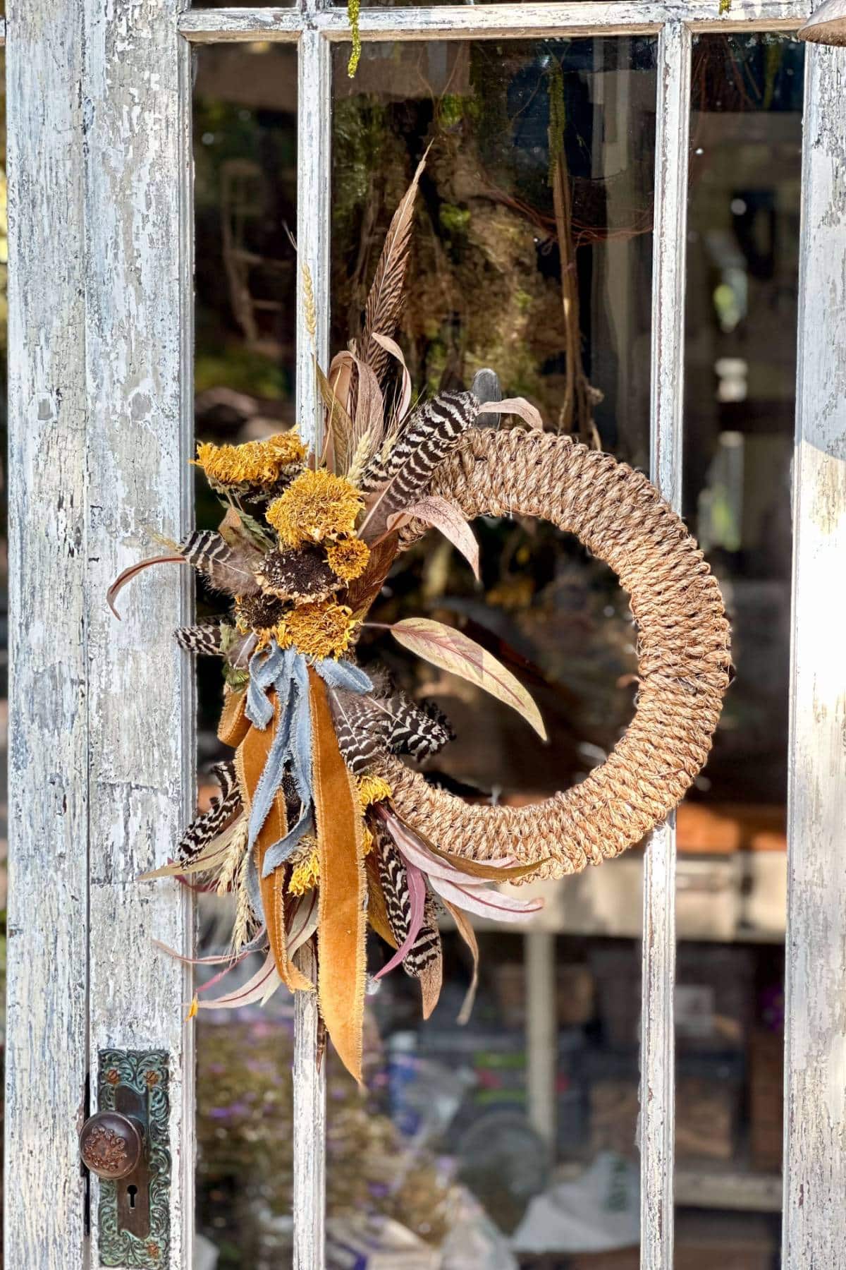 A rustic door with peeling white paint features a decorative wreath made of pinecones, feathers, and yellow and brown dried flowers. The wreath is hanging on the glass pane, adding a touch of natural, autumnal charm to the weathered door.