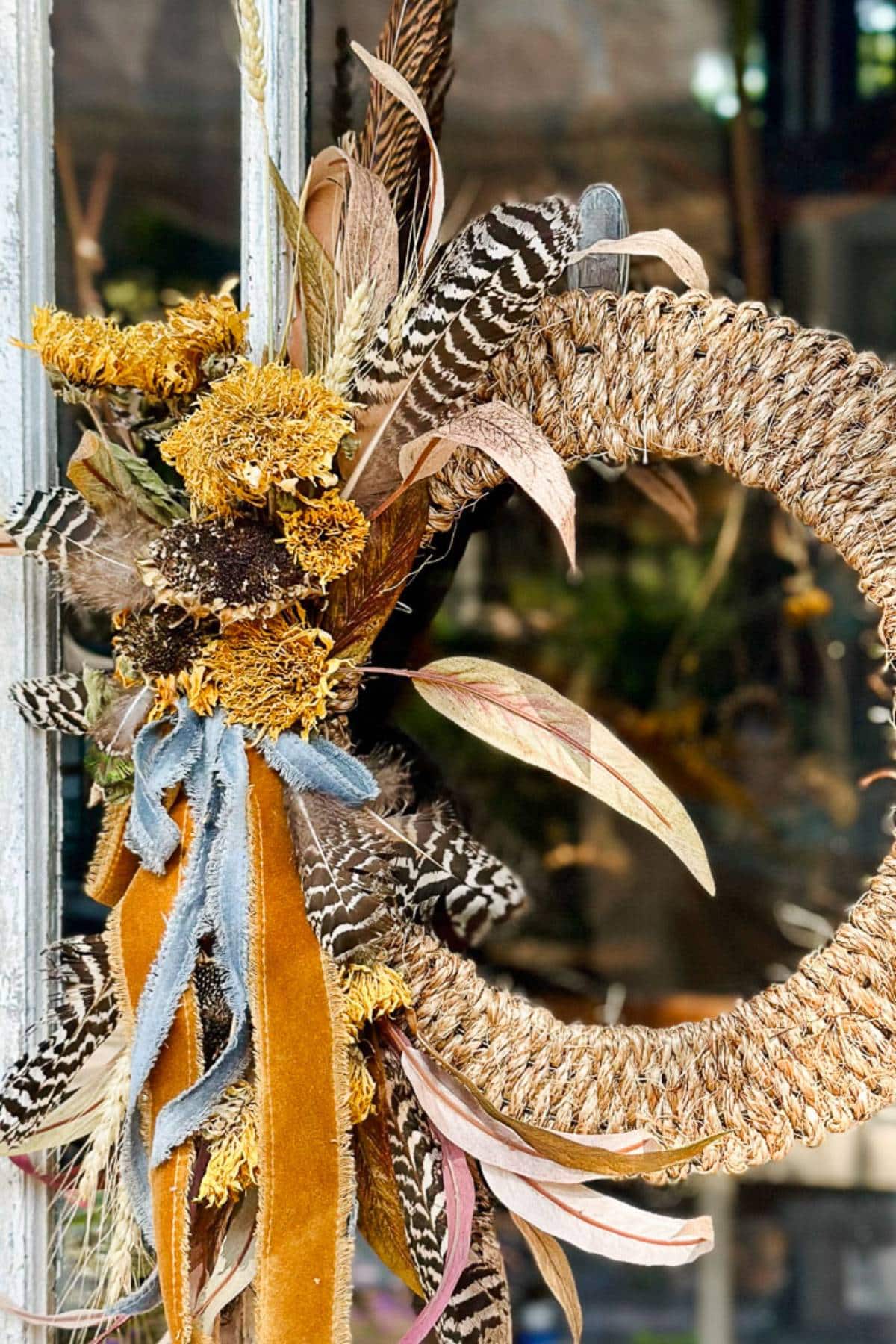 A rustic wreath made of woven rope and decorated with a mix of dried flowers, feathers, and leaves hangs on a door. The colors include shades of yellow, brown, and green, with an orange ribbon adding a touch of warmth to the arrangement.