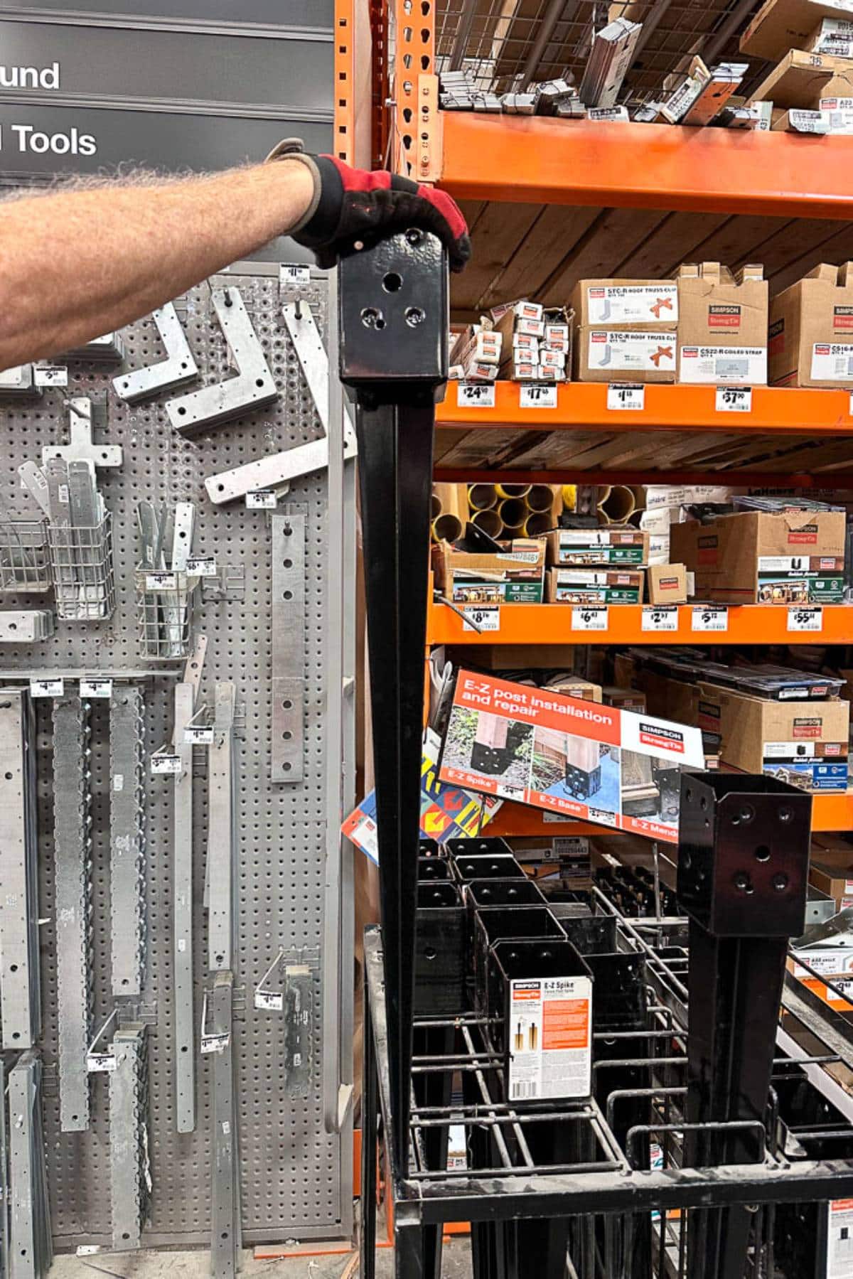 A person wearing a glove is holding up a black metal post bracket in a hardware store aisle. The background features orange shelving with various construction materials and accessories, including metal brackets and boxes on display.