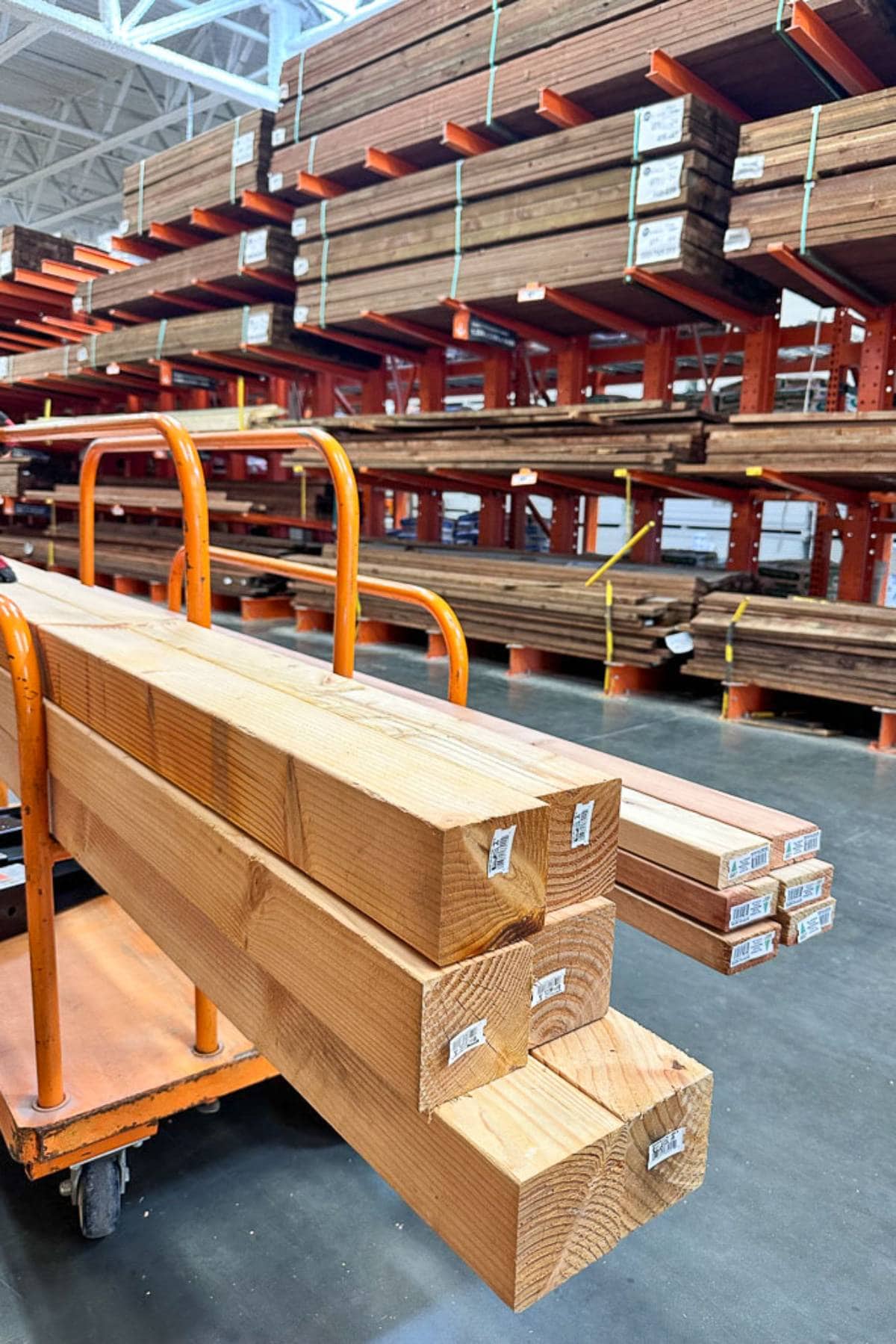 Stacks of wooden planks are displayed on metal shelves in a hardware store. Several large rectangular wooden beams are placed on an orange cart in the foreground. The scene is well-lit, showcasing the organized layout of the lumber section.