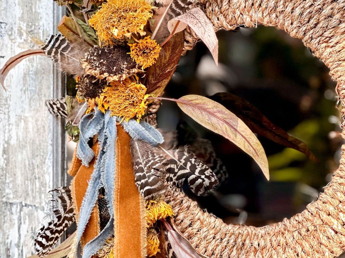 A close-up of a rustic wreath made from dried flowers, natural feathers, and brown and orange ribbons. The DIY rope wreath features a circular, woven texture and is attached to a weathered surface, contributing to the natural and vintage aesthetic perfect for the fall season.