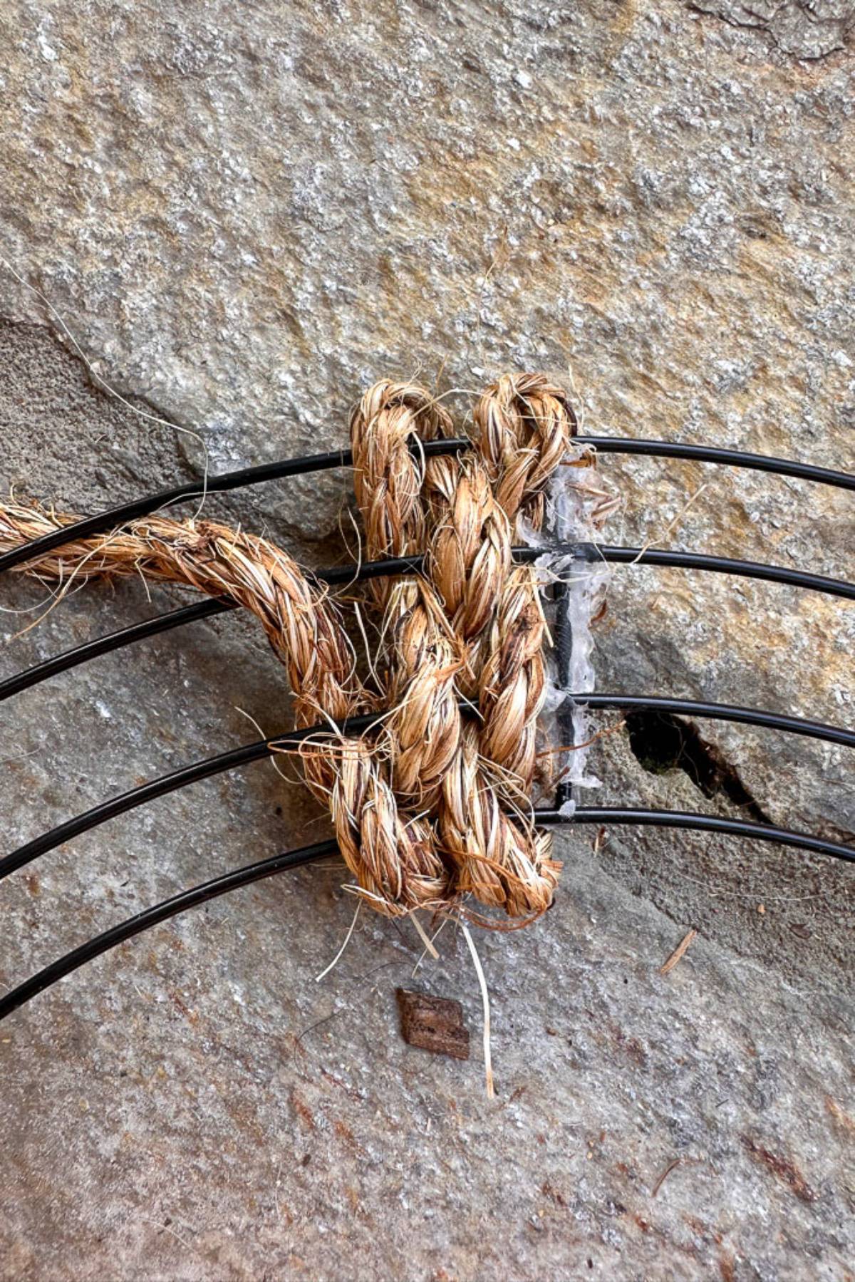 Close-up of a knot made with thick, fibrous rope possibly holding together several black wires, all set against a stone or concrete surface with visible texture.