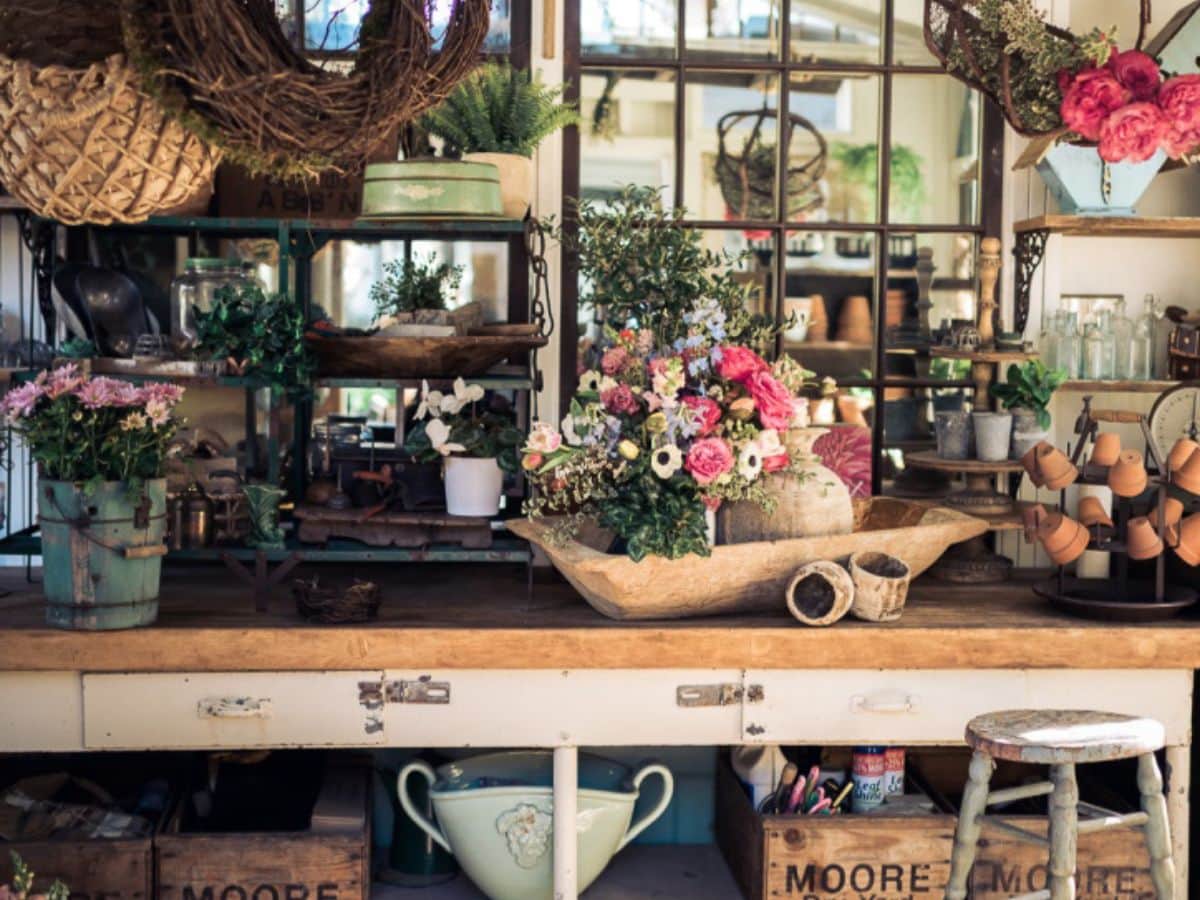 A rustic, cluttered workspace is adorned with various potted plants and flowers. The wooden desk holds gardening tools, baskets, and decorative items. Shelves and a mirror in the background reflect the cozy, charming atmosphere, offering thrifting tips for beginners amid the stool and wooden crates in the foreground.