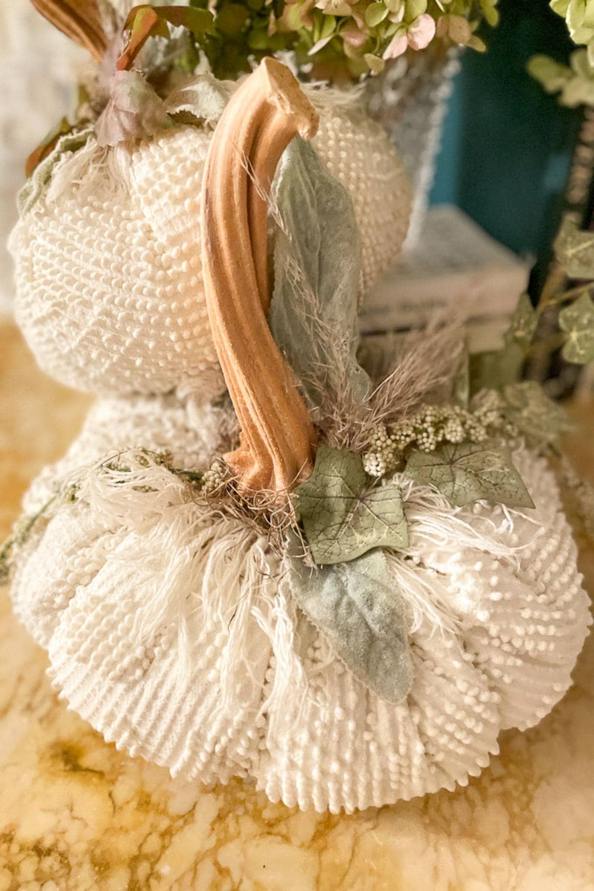 Two white fabric pumpkins sit on a marble surface. Each pumpkin is adorned with textured beads and a tan-brown stem, with light green fabric leaves and a decorative flower crown around the base of the stem. The background includes a glimpse of foliage and a book.