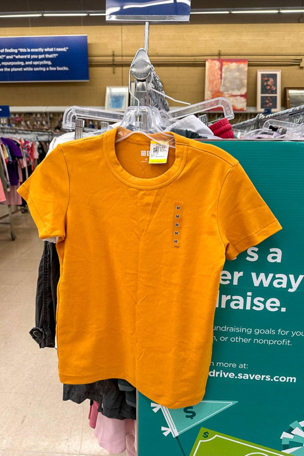 An orange short-sleeve t-shirt hangs on a clear plastic hanger in a thrift store. The shirt is displayed against a backdrop of other clothes and a green sign promoting fundraising through secondhand clothing drives.