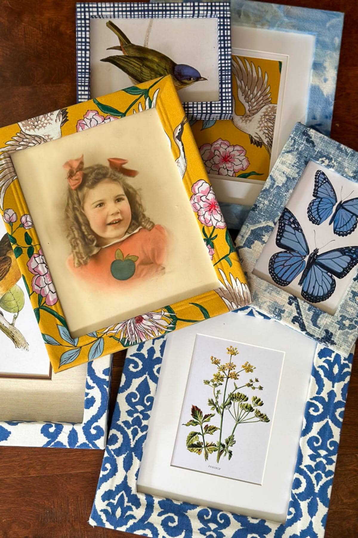 A collection of colorful picture frames lies on a wooden surface. The frames feature a mix of patterns, including florals and geometric designs. They showcase art pieces such as a portrait of a young girl with bows in her hair, birds, butterflies, and botanical illustrations.