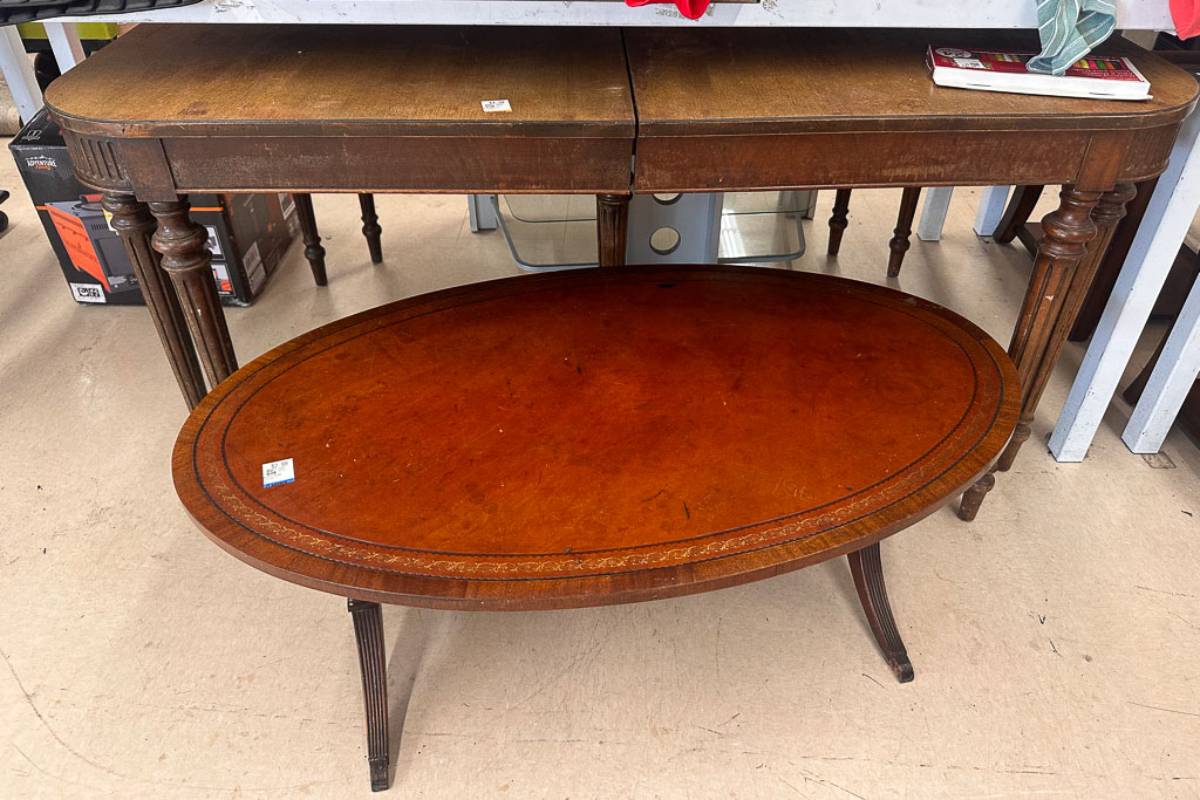 Two wooden tables for sale: a rectangular table with turned legs in the background and an oval coffee table with a reddish-brown finish and curved legs in the foreground. The tables stand on a light-colored floor.