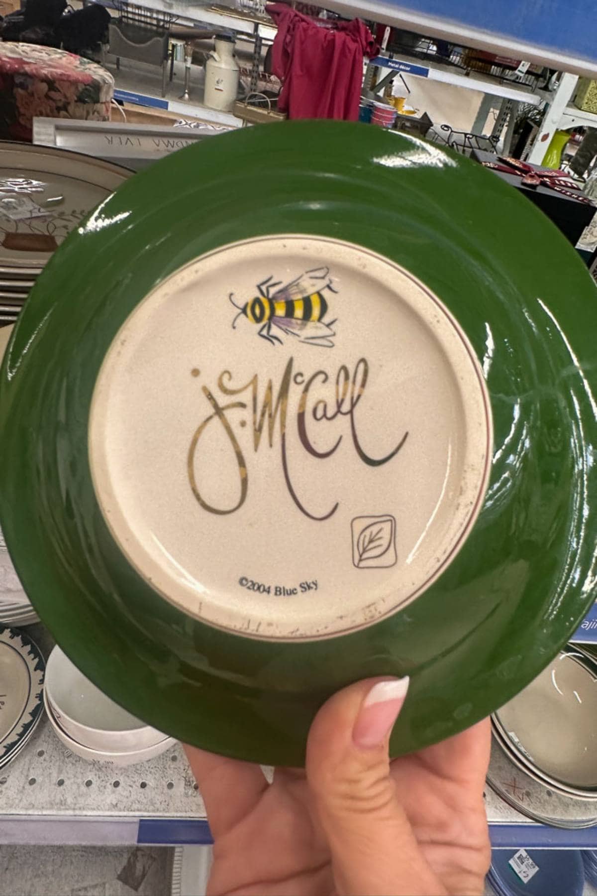 A white plate with a green rim is held face-down. The bottom of the plate features a bee illustration and the signature "J. McCall." The text "©2004 Blue Sky" and a small leaf design are also visible. Various dishes are displayed in the background.