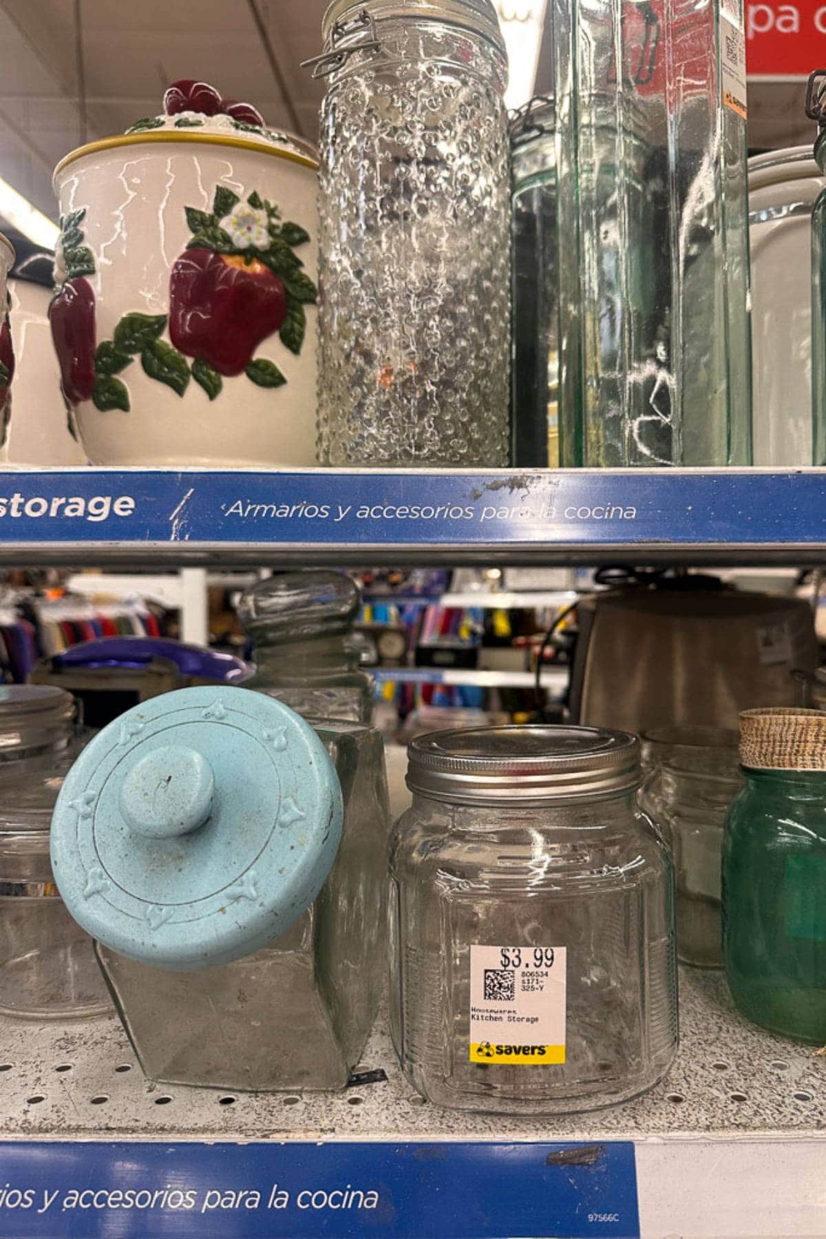 A thrift store shelf displays various glass storage jars, including a tall textured jar, a jar with a turquoise lid, and a large jar with a small price tag of $3.99. There is also a ceramic container with an apple design and additional kitchen items in the background.