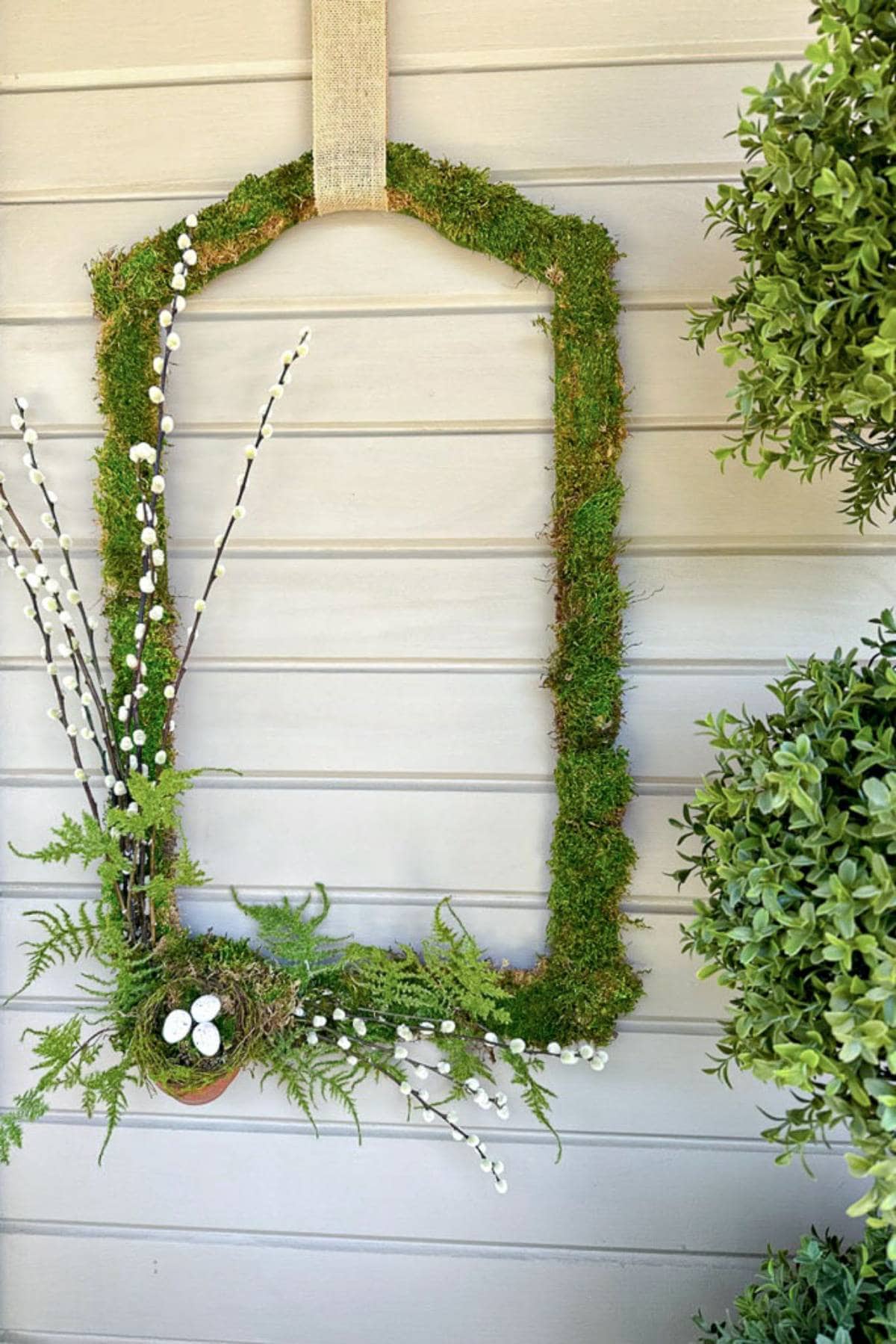 A unique rectangular wreath covered in green moss is hanging on a light gray wall. It is decoratively adorned with white pussy willow branches and ferns at the bottom, creating a natural and rustic appearance. Two green shrubs frame the sides of the image.