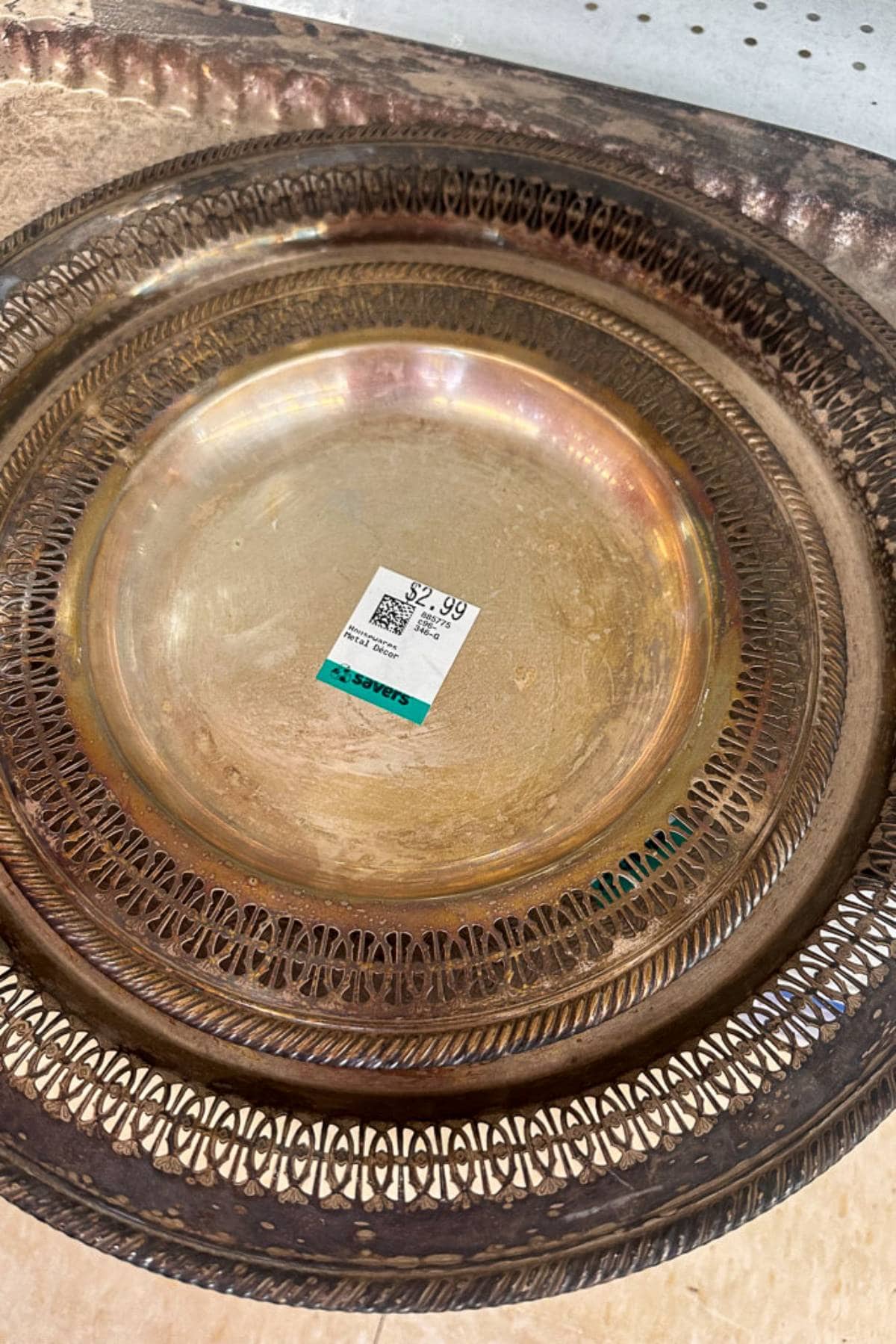 A close-up photo of three intricately designed, silver-colored plates stacked together on a store shelf. The topmost plate has a price tag displaying $1.99. The edges of the plates feature ornamental patterns.