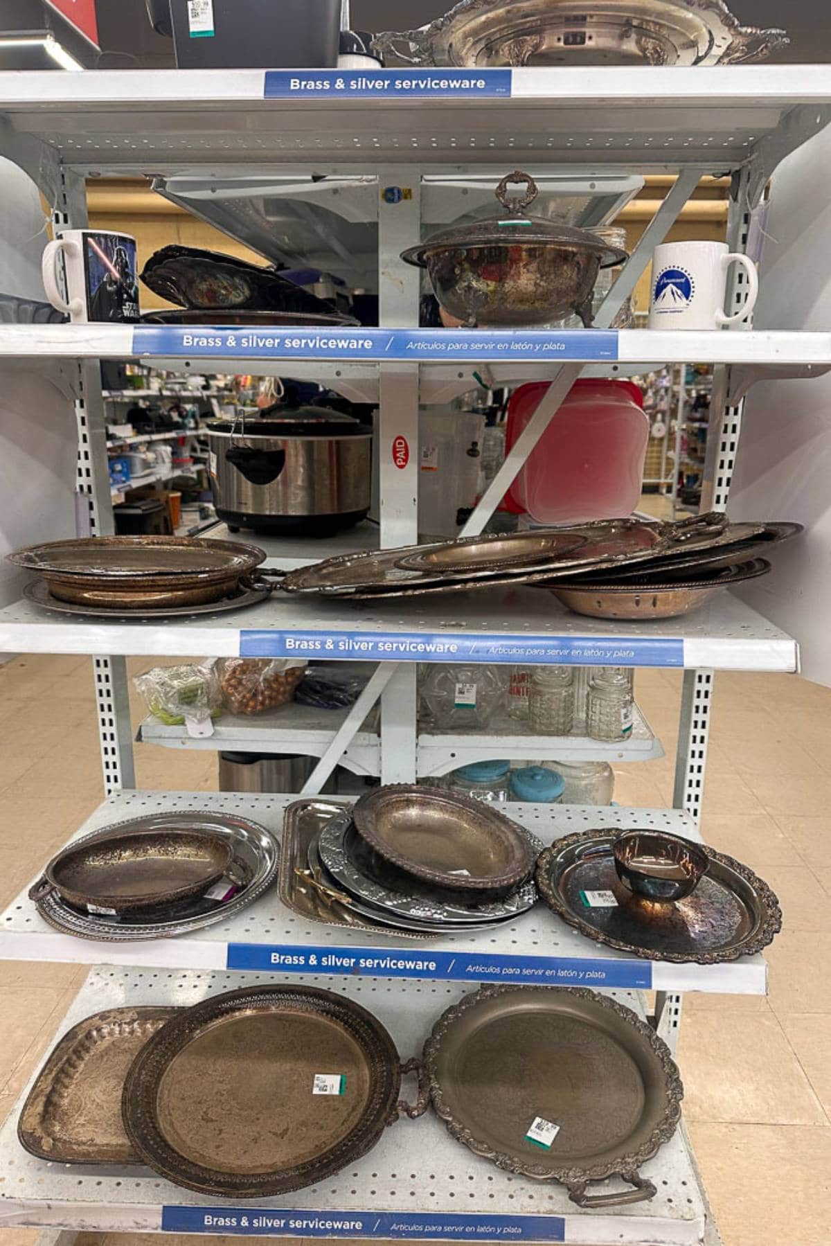 Shelves displaying various brass and silver serviceware items, including trays, bowls, mugs, and platters. The serviceware appears tarnished and vintage. Shelves are labeled "Brass & Silver Serviceware" and the background hints at a store or thrift shop setting.