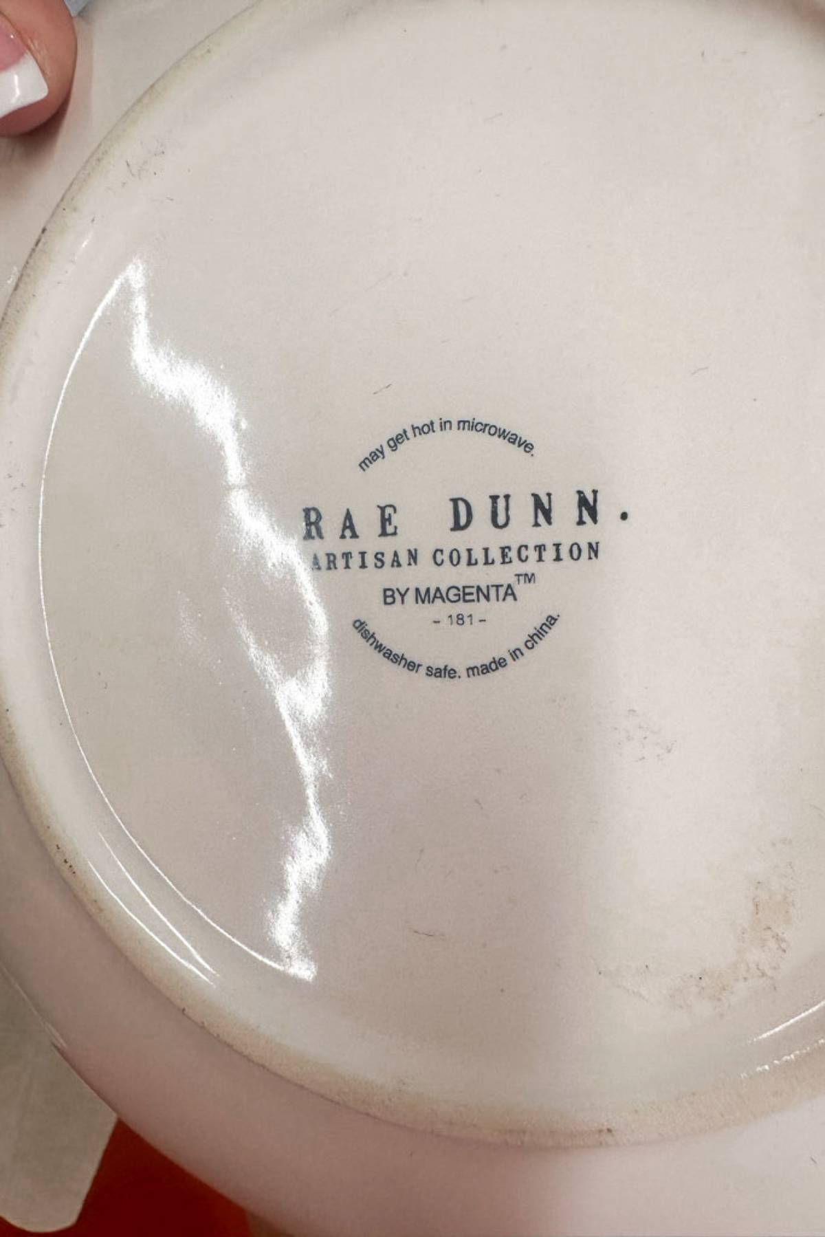 The bottom of a ceramic dish is shown with the text: "RAE DUNN ARTISAN COLLECTION BY MAGENTA ™ 181- dishwasher safe, made in china" along with a note that says, "may get hot in microwave". The surface appears smooth and slightly reflective.