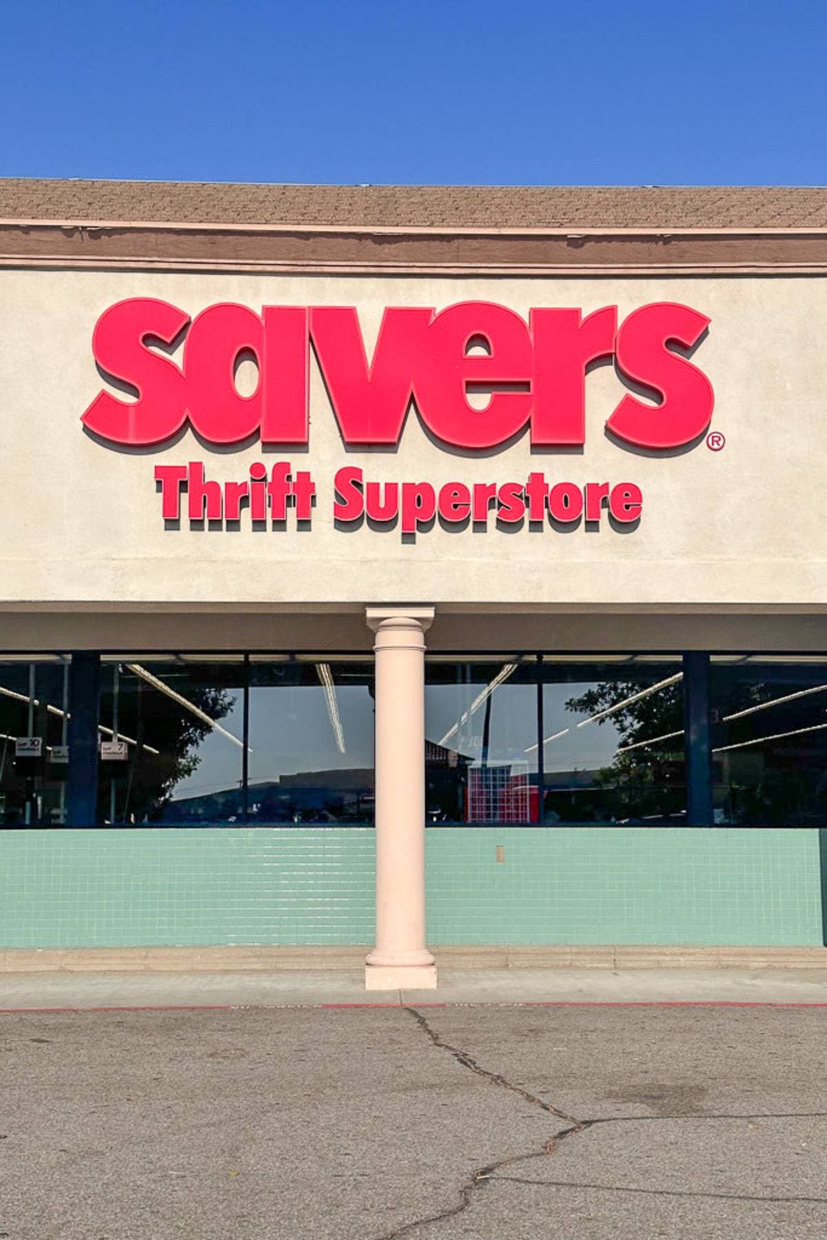 Image of the front entrance of a Savers Thrift Superstore. The storefront features a large red Savers sign above the words "Thrift Superstore" on a beige background, with a single beige column in front of the glass windows and doors. The parking lot is visible.