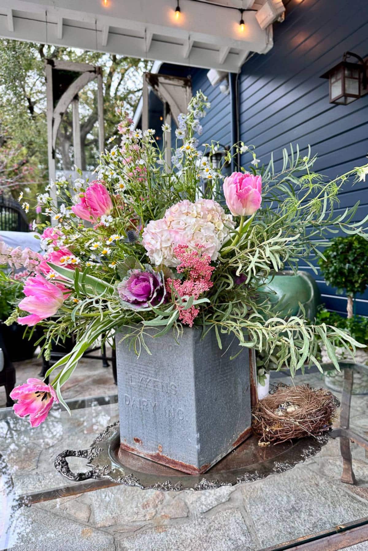 A rustic floral arrangement in a vintage metal container is displayed on a glass table. The bouquet features pink tulips, white hydrangeas, small daisies, and greenery. A small empty bird's nest is placed nearby on the table.