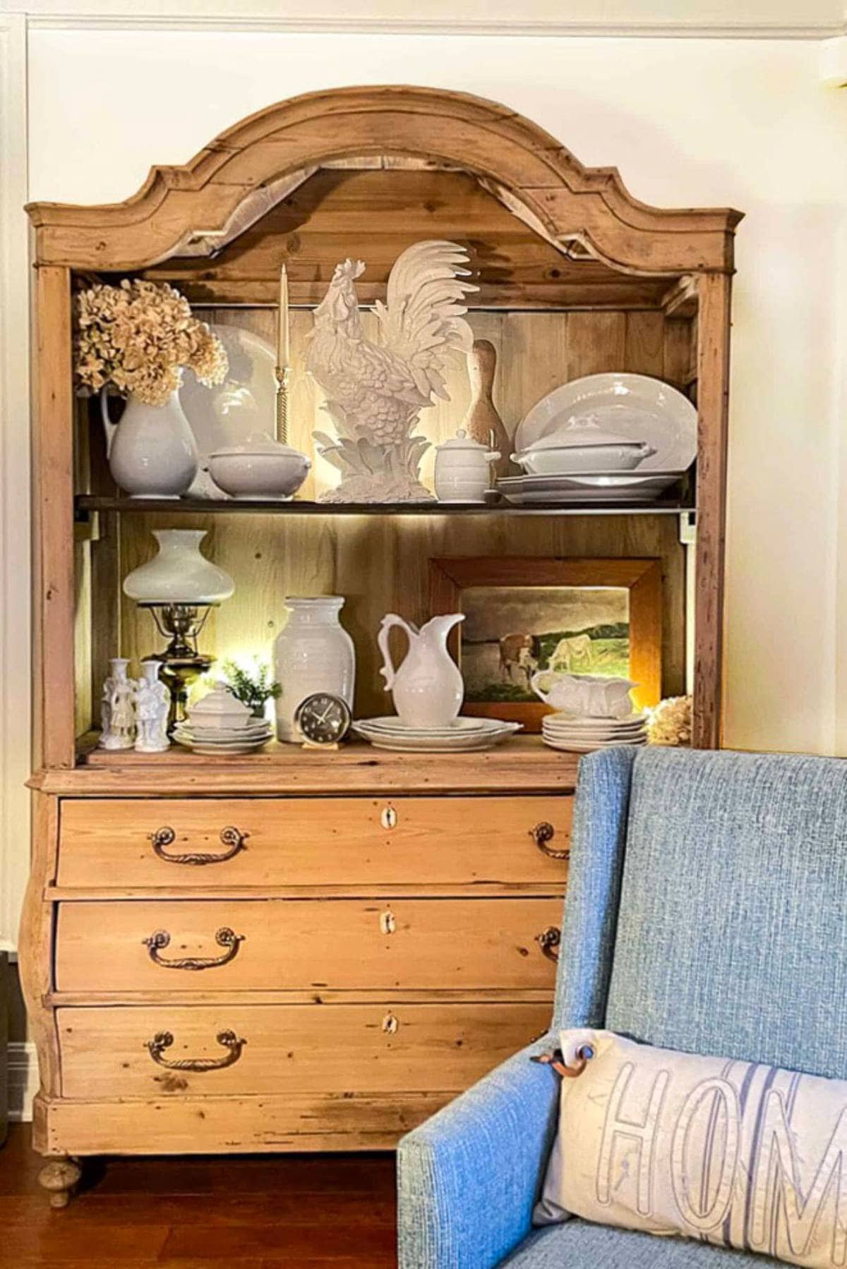 A wooden hutch with ornate detailing displaying white ceramic dishes, a large rooster statue, and dried flowers is positioned against a wall. Below it is a three-drawer cabinet. To the right, a blue armchair with a pillow that reads "Home" is partially visible.