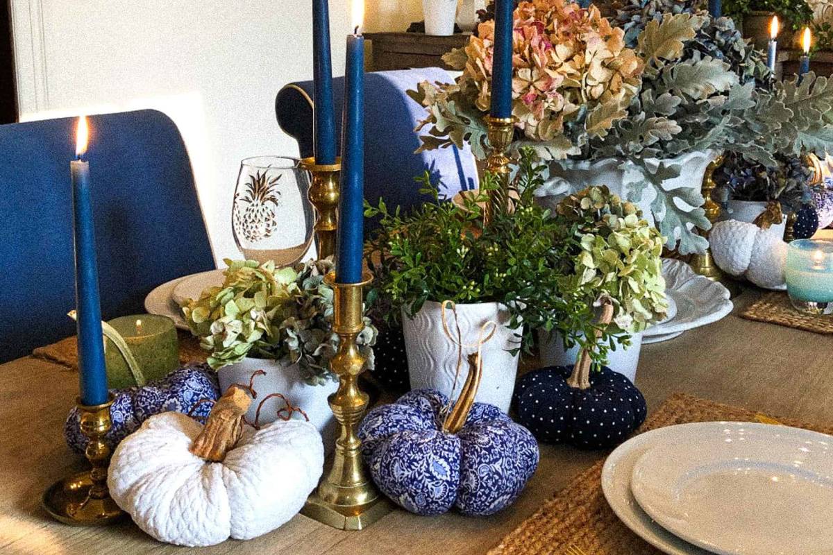 A dining table decorated for fall with blue and white fabric pumpkins, brass candlesticks holding blue candles, potted plants, autumnal foliage, and white plates set on woven placemats. The candles are lit, creating a cozy and warm atmosphere.