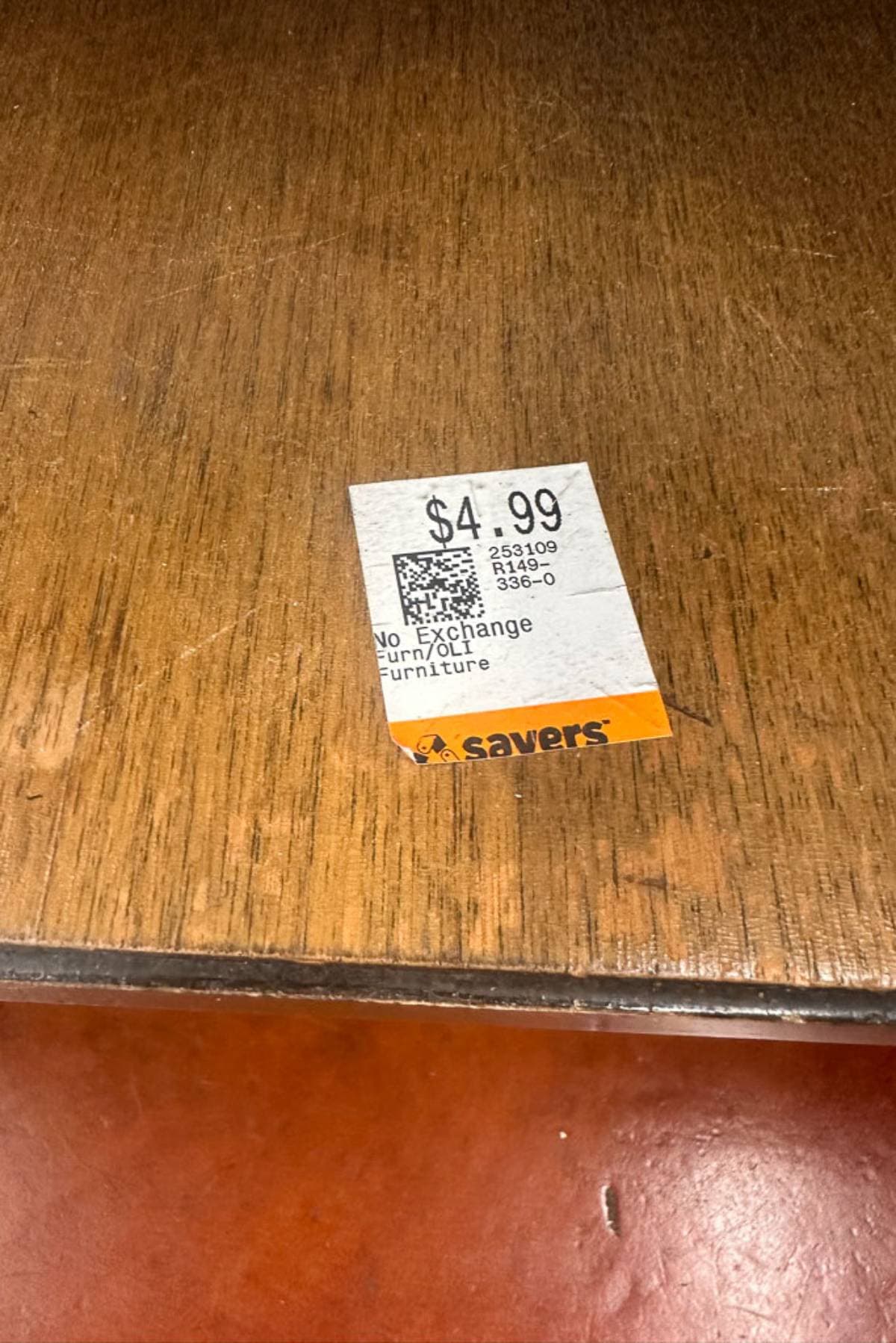 A white price tag from Savers placed on a wooden surface. The tag indicates a price of $4.99 and includes a barcode and some text that mentions "No Exchange" and "Furniture".