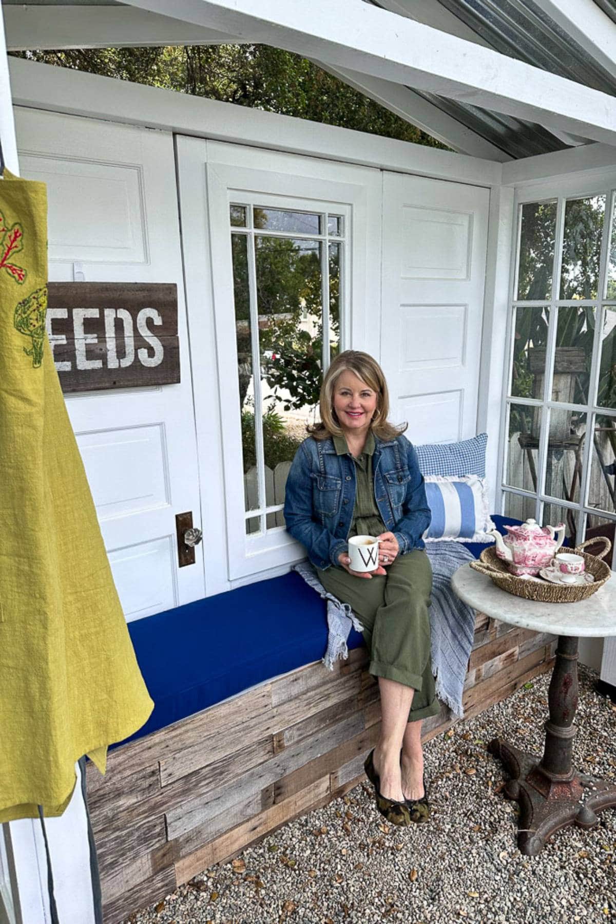 A woman sits on a cushioned bench in a cozy garden shed, smiling while holding a white mug with the letter "W" on it. She wears a denim jacket and green outfit, surrounded by pillows. A table with teacups and a teapot is nearby. A "SEEDS" sign hangs on the wall.