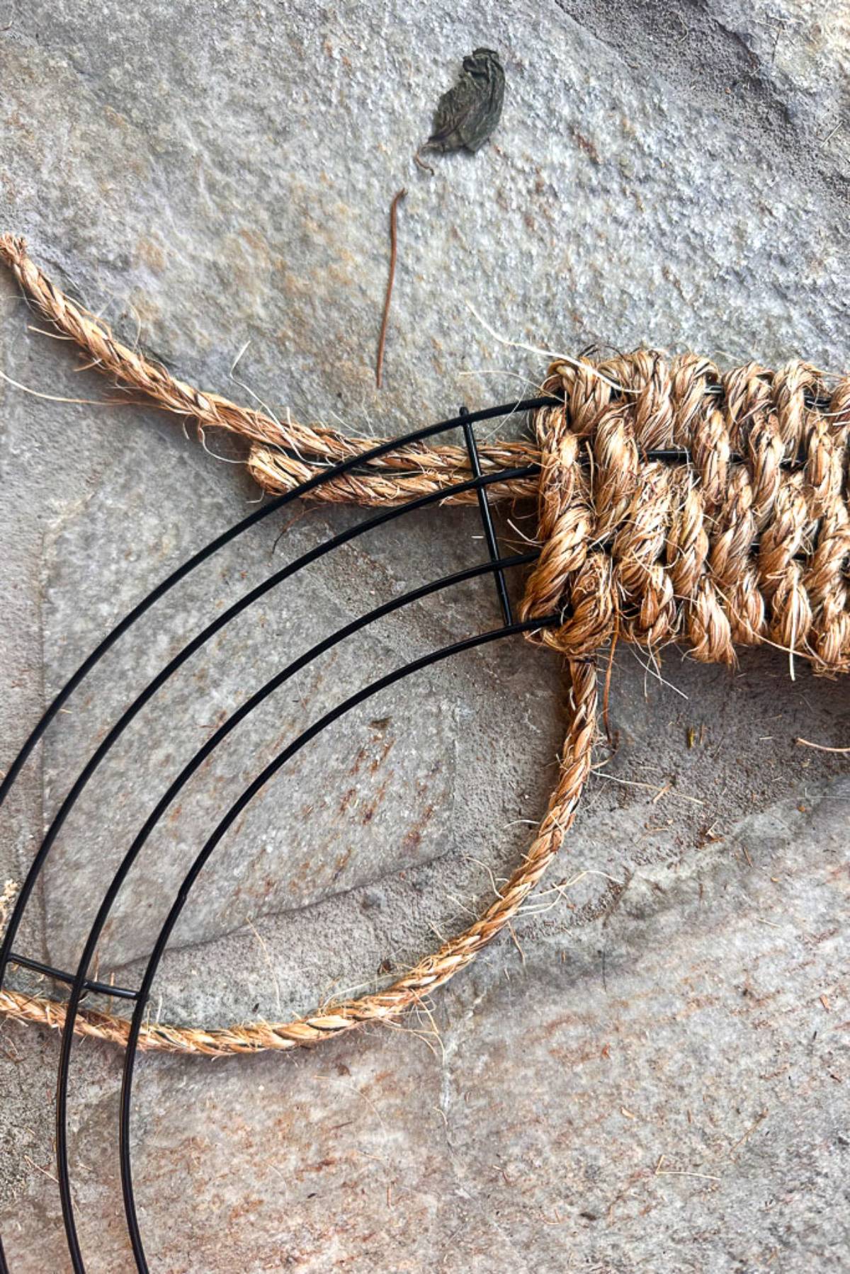 Close-up of a partially completed craft project using black metal rings and coarse brown rope. The rope is wrapped tightly around the metal rings and tied off, forming a design on a textured stone surface. A dried leaf is visible in the background.