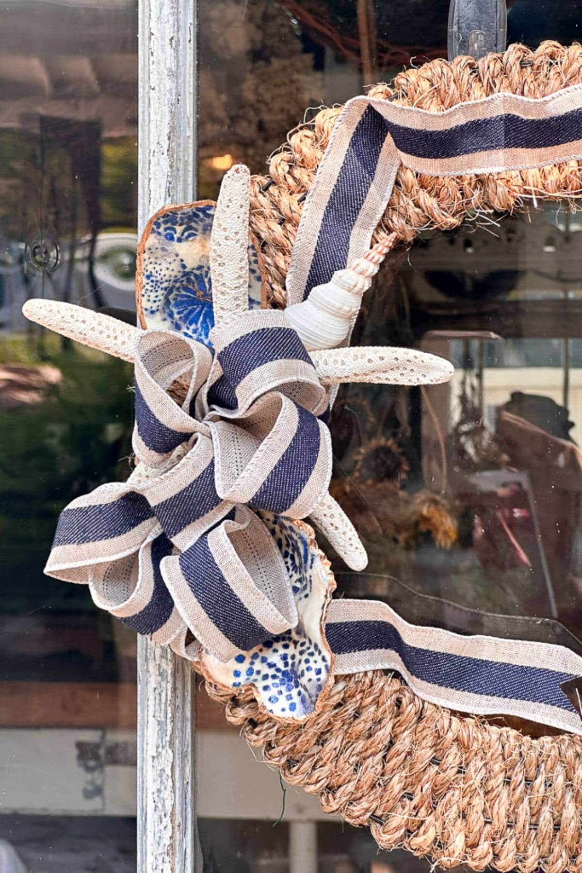 A decorative wreath made of natural materials is adorned with a large seashell, a starfish, and a ribbon with blue and white stripes. The wreath is hanging on a glass door, reflecting the outdoors, with a blurred background.