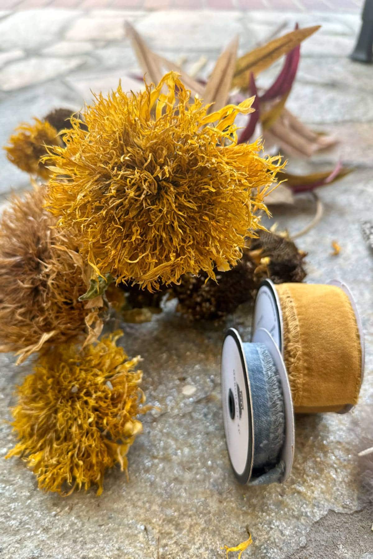 A collection of dried yellow flowers rests on a stone surface. Beside the flowers, two spools of ribbon in yellow and grey are placed next to each other. The background shows additional natural elements, creating a rustic and autumnal feel.