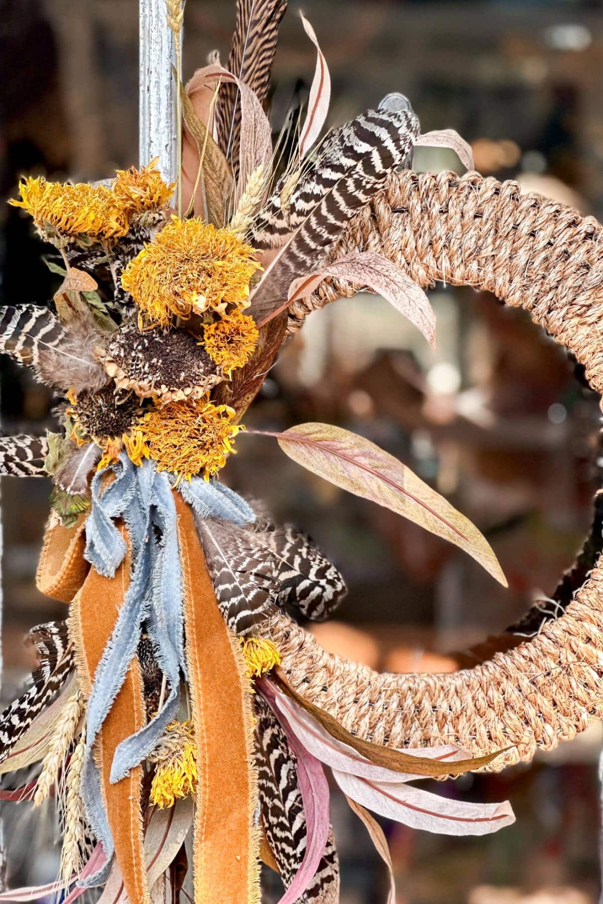 A close-up of a decorative wreath adorned with various dried flowers, leaves, feathers, and textured ribbons. The wreath is made from a twine material and the background is blurred, focusing attention on the intricate details of the arrangement.