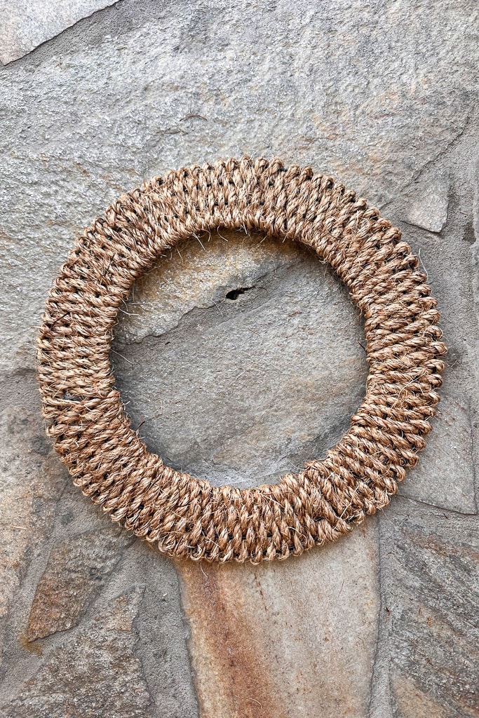 A circular woven DIY Rope Wreath made of natural, textured rope is displayed on a stone surface. The wreath consists of tightly wound fibers creating a rustic, handmade look, perfect for the Fall Season. The stone background enhances its earthy, natural aesthetic.