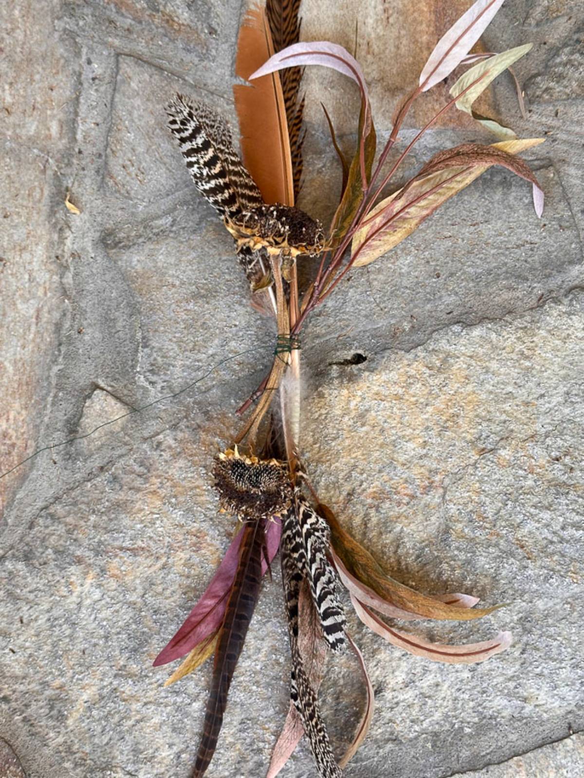 A decorative bundle hangs on a stone wall. The arrangement includes a mix of brown, black, and white striped feathers and dried leaves in shades of brown and red. The feathers and leaves are tied together with a thin piece of green string.