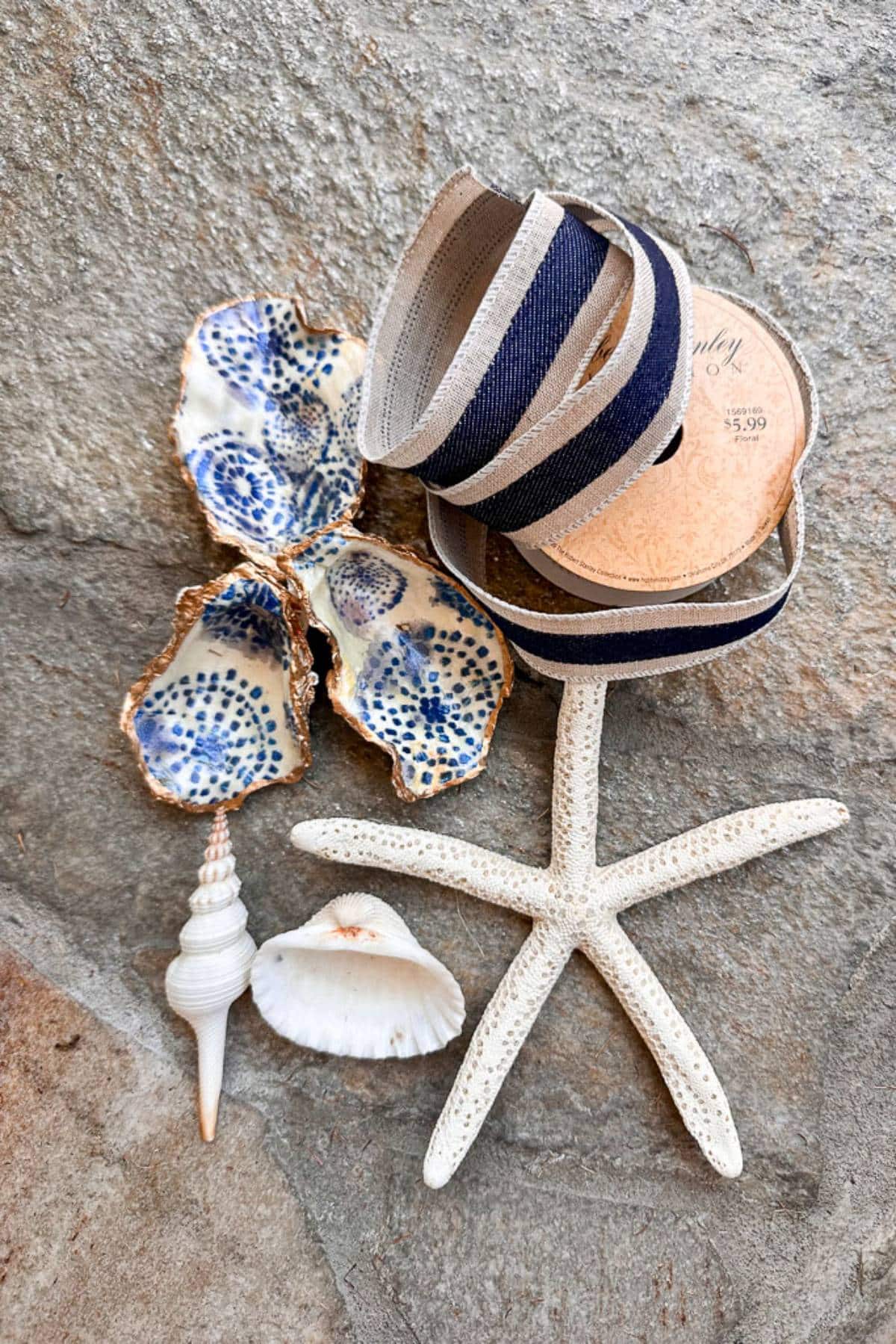 A textured surface holds a starfish, seashells, two blue and white oyster shells, and a roll of navy and beige ribbon. The starfish is centered at the bottom, and the ribbon is coiled on a cardboard spool labeled with "5.99".