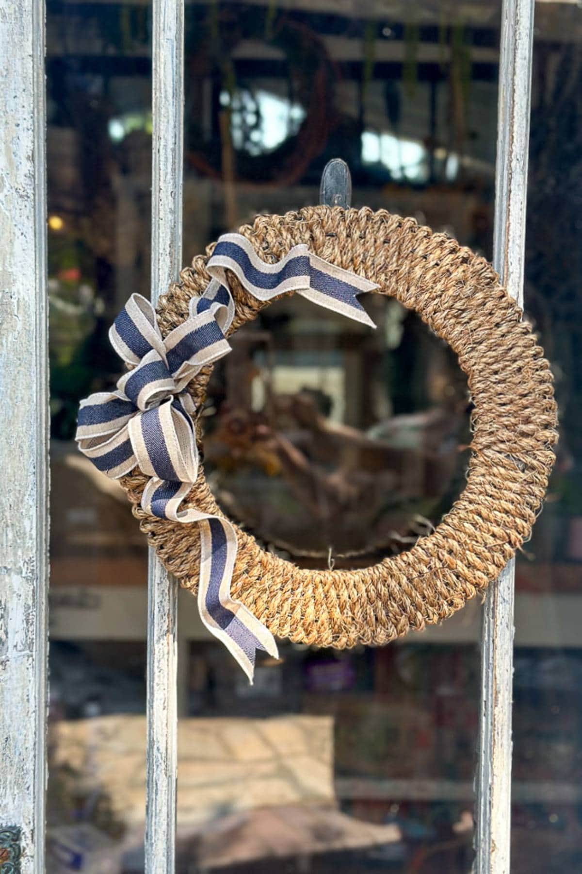 A rustic wreath made of looped natural rope is hung on a windowpaned door. The wreath is adorned with a decorative bow in black and white stripes, adding a touch of charm to the quaint door. The background features a blurred indoor space.