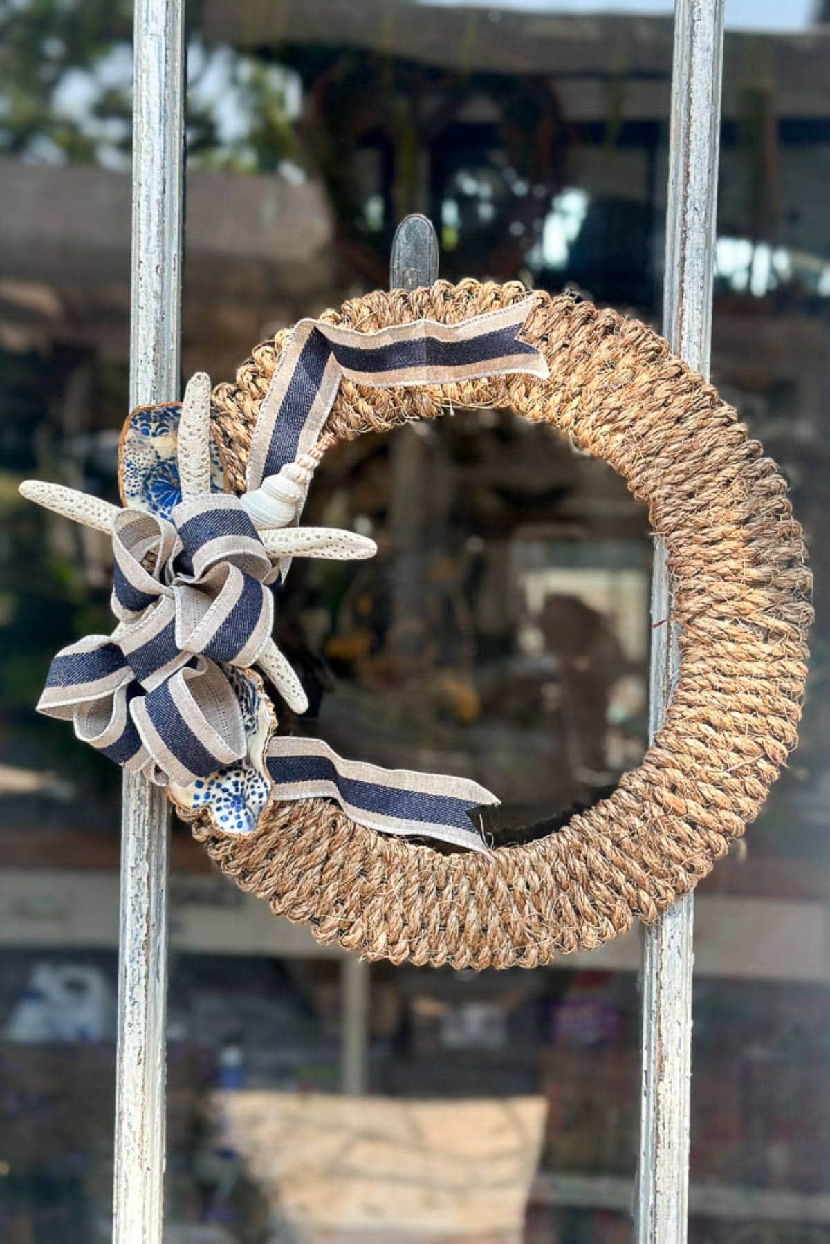 A decorative wreath made of thick, braided natural fibers hanging on an old wooden door. It is adorned with a cluster of seashells and a striped ribbon bow in navy blue and white on the left side. The background shows blurry outdoor elements through the glass.