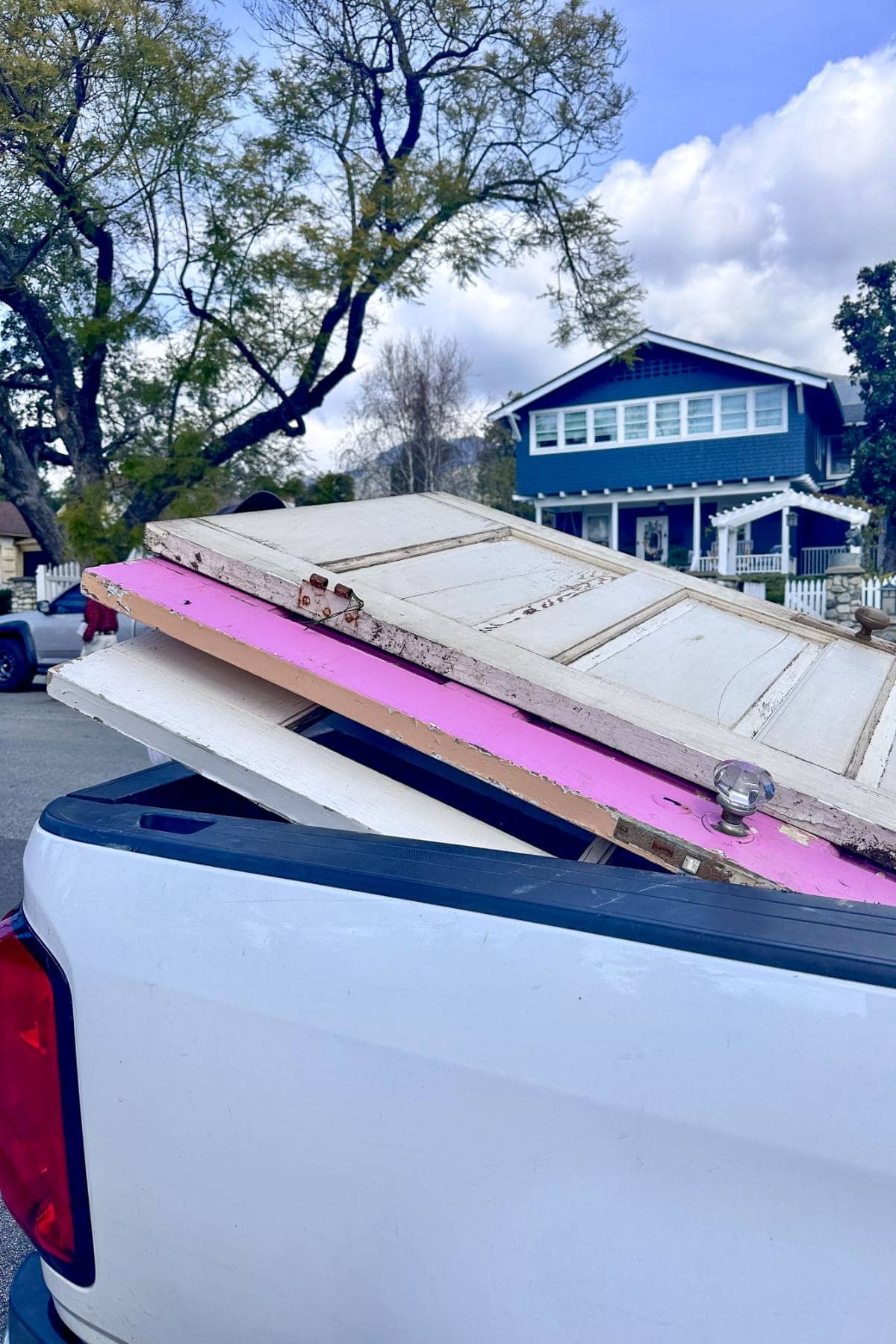 A white pickup truck carrying old doors, including a white one and a pink one, in its bed. In the background, there's a blue two-story house with white trim, trees, and a partly cloudy sky.