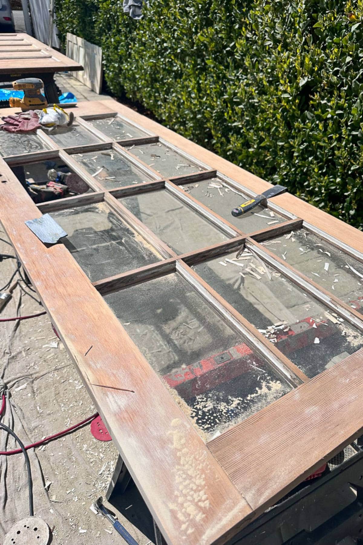 A wooden door with multiple glass panes is lying flat on a tarp-covered surface outdoors. Various tools and materials are scattered on and around the door, and sawdust is visible. Green hedges line the background.