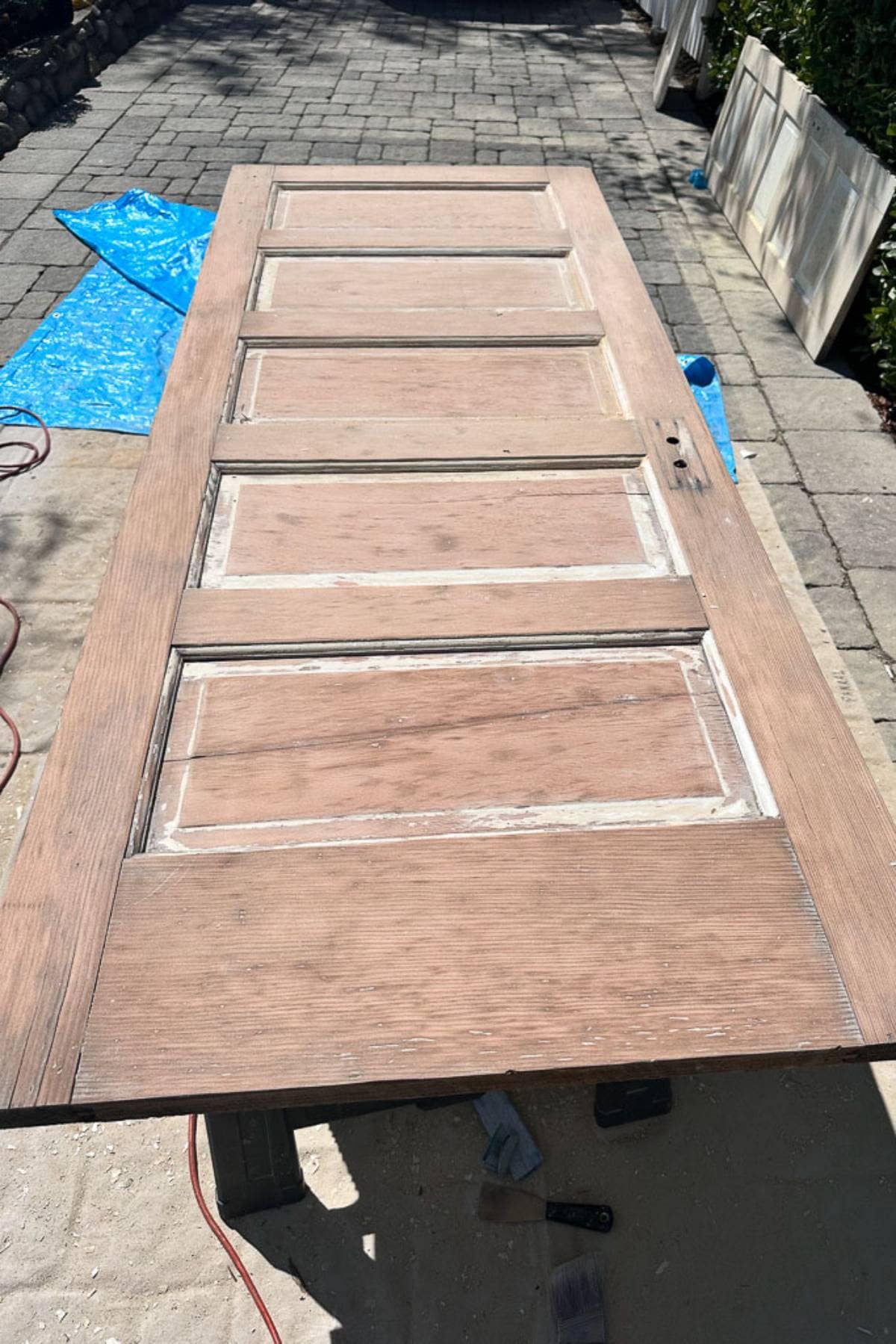 A wooden door with five panels lies on sawhorses outdoors. The door appears to be in the process of being sanded or refurbished, with tools and a blue tarp visible in the background on a paved surface.