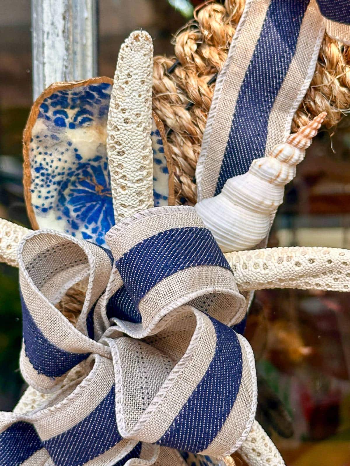 A decorative arrangement featuring a starfish adorned with a blue and beige patterned ribbon, a small white seashell, and strands of lace and woven material. The backdrop includes a piece of ceramic with blue splashes, adding a nautical touch.