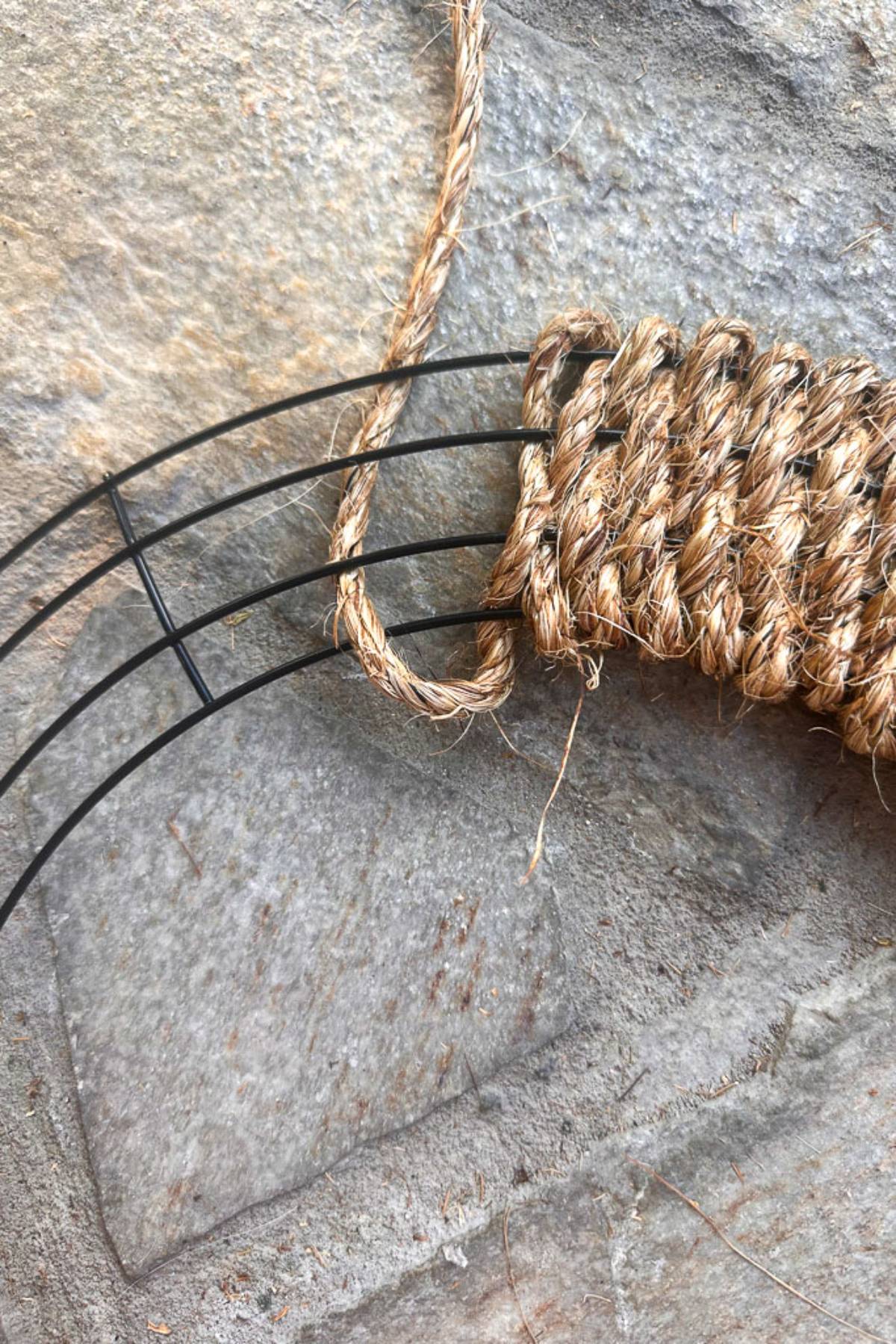 Close-up view of a craft project, showing a wire frame being wrapped with thick natural twine. The wrappings are partially completed and the project is placed against a stone surface. The twine's fibers and the wire frame's structure are visible in detail.