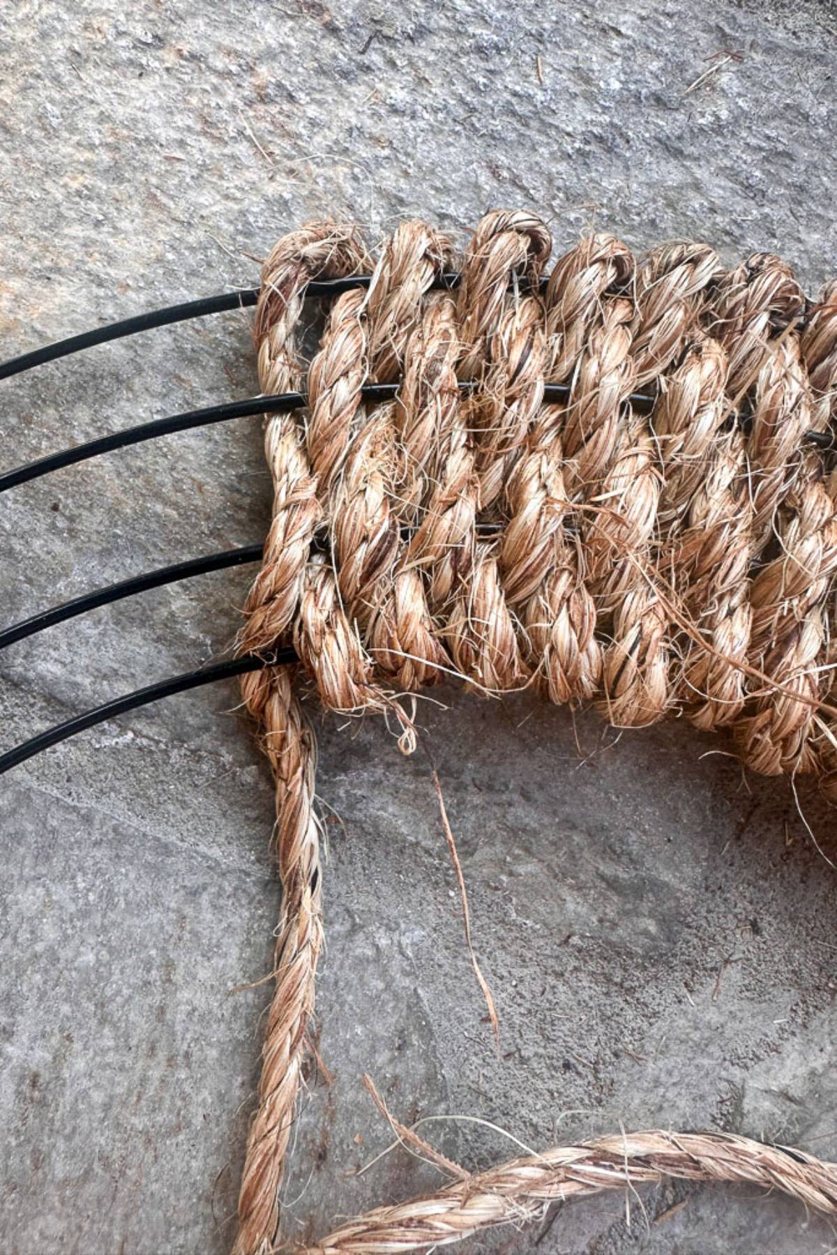 Close-up of a weaving project in progress. Natural fiber ropes are interwoven with black plastic cords on a gray stone surface, showing intricate patterns and knots. One loose end of the fiber rope is visible, indicating the project is still being worked on.