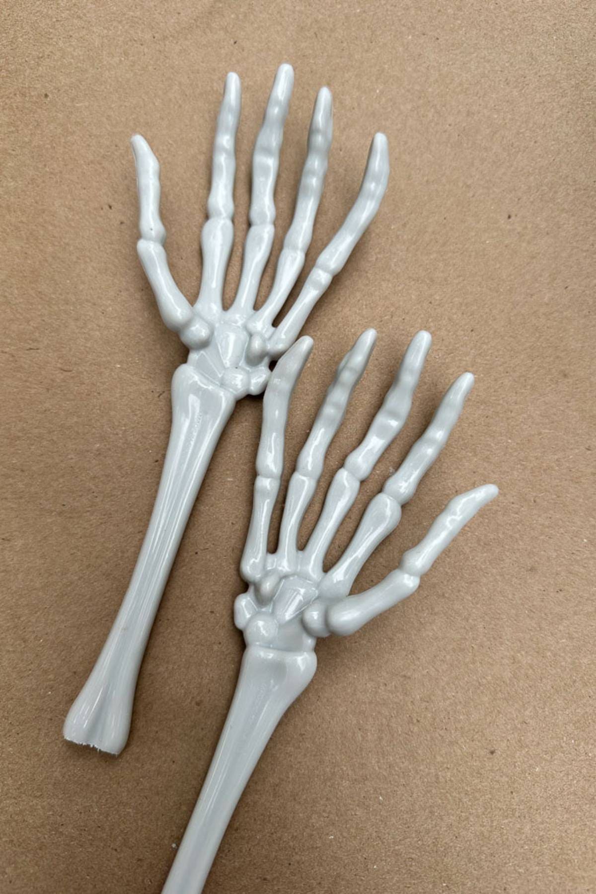 Two plastic skeleton hand-shaped utensils are laid on a brown surface. Featuring intricate bone detailing, these hands resemble human skeletal hands, making them perfect for setting the stage at your Halloween table or even as quirky DIY Halloween place card holders.