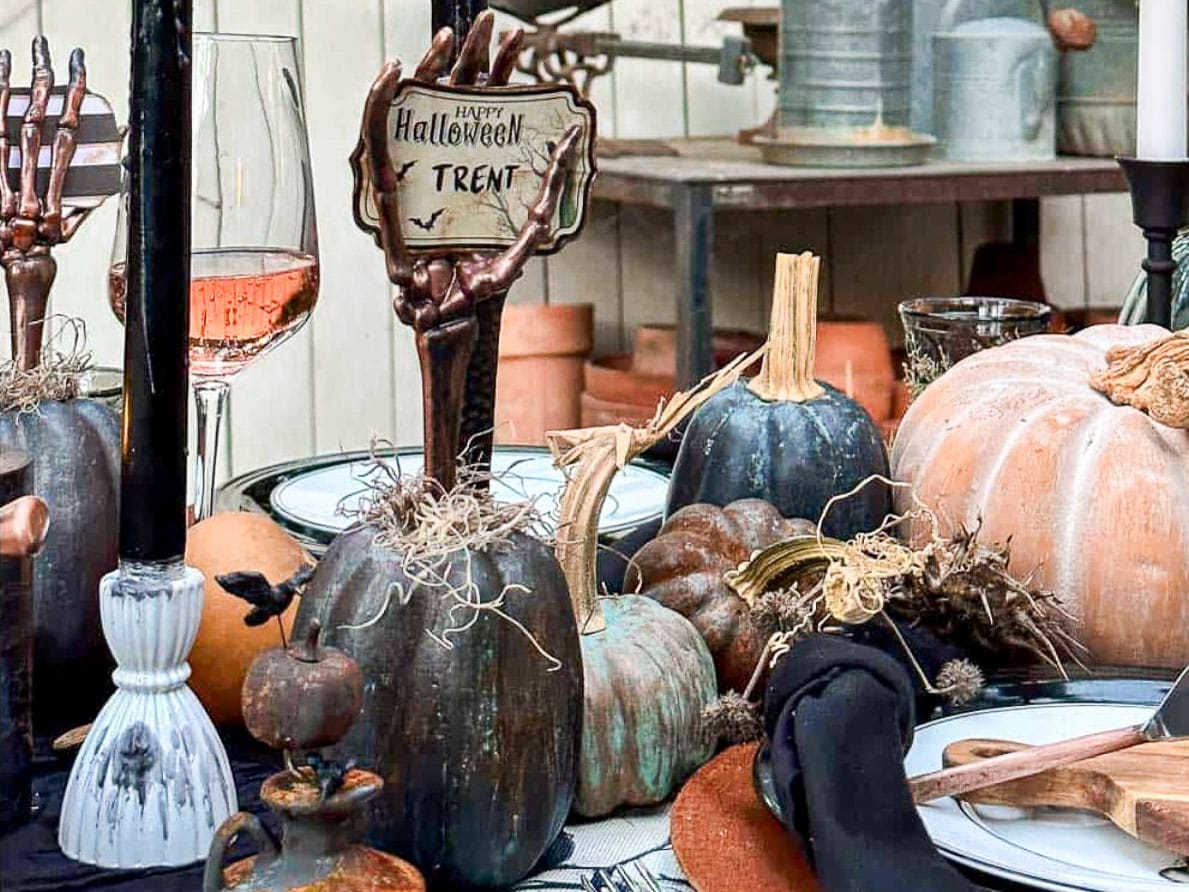 A spooky Halloween-themed table setting featuring black pumpkins, a sign that reads "Happy Halloween Trent," candles, a glass of wine, and rustic decorations. The table includes black and white plates, cutlery, small pumpkins, props, and DIY Halloween place card holders with free printables for an extra special touch.