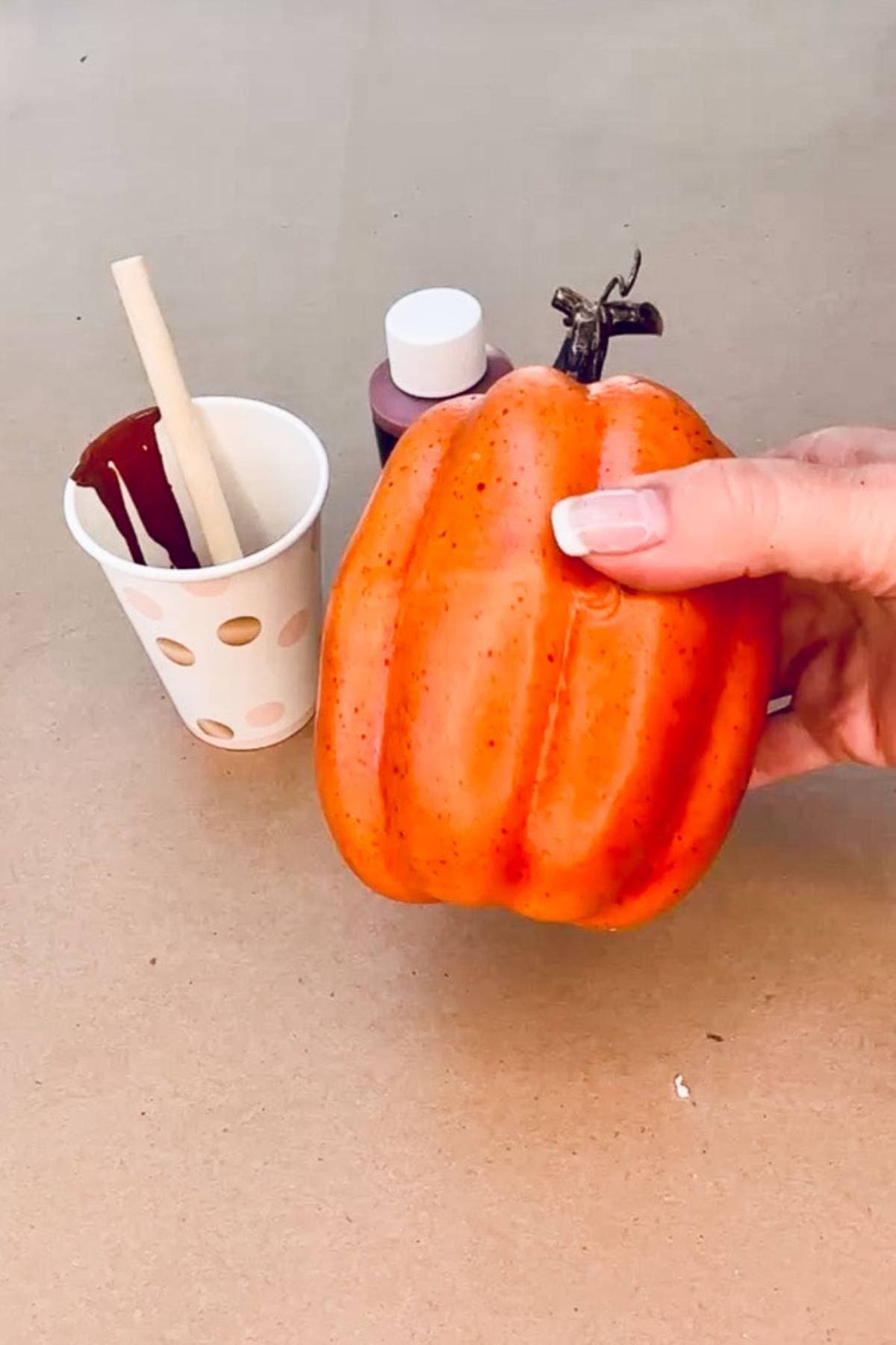 A hand holds a small orange pumpkin, perfect for crafting DIY Halloween place card holders, while a paper cup with a paintbrush and brown paint and a bottle of paint rest on the Halloween table.